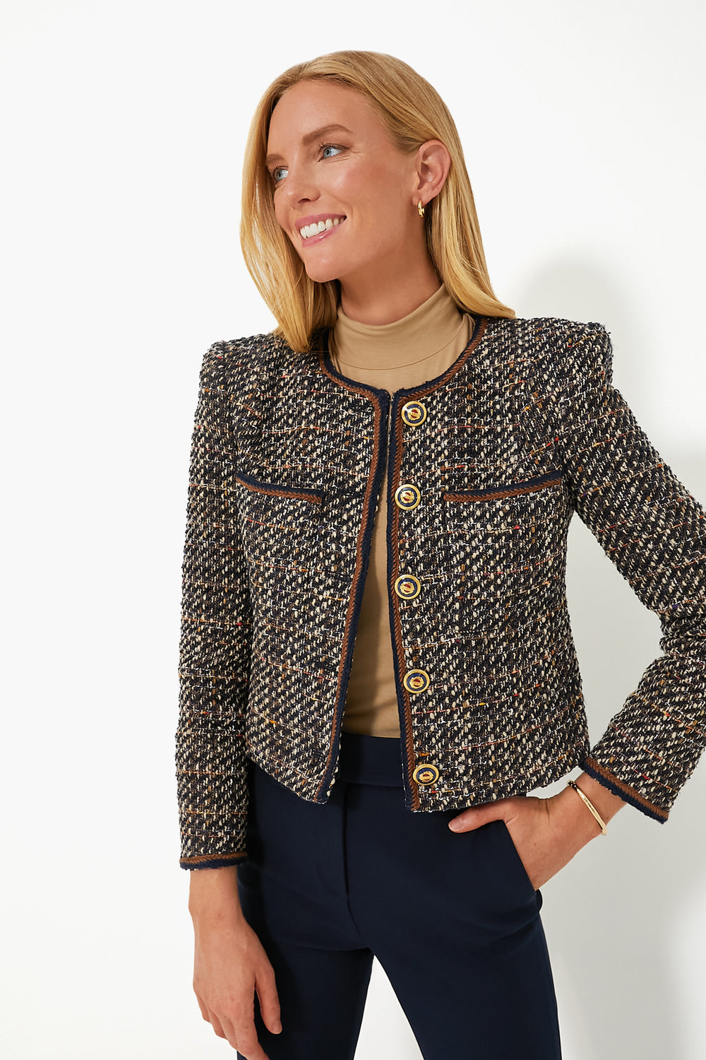 A woman smiles with one hand on her hip, wearing a Veronica Beard boucle tweed jacket with gold buttons over a beige turtleneck and dark pants.