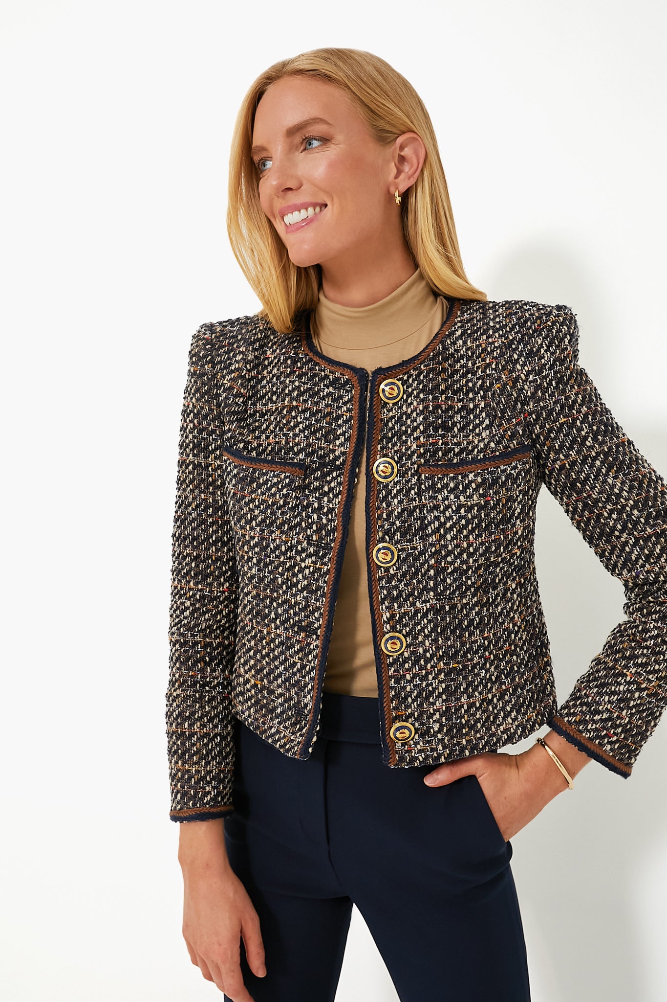 A woman smiles with one hand on her hip, wearing a Veronica Beard boucle tweed jacket with gold buttons over a beige turtleneck and dark pants.