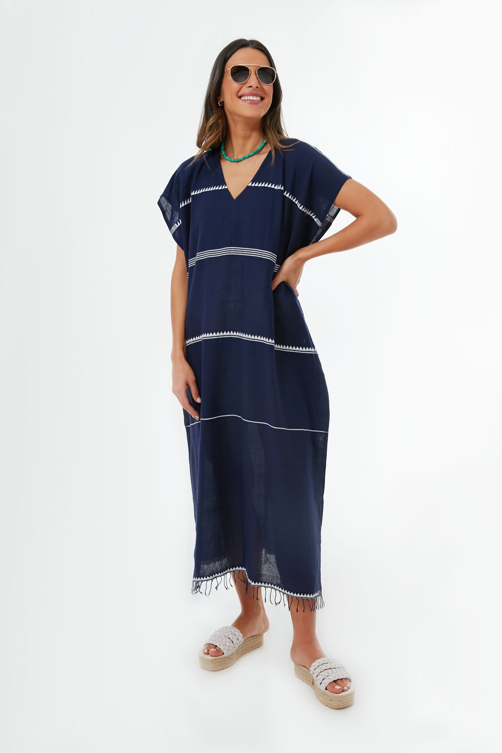 A woman wears sunglasses and a navy lemlem Nunu Classic Caftan resort dress with white trim, green necklace, and white platform sandals—handmade in Ethiopia.