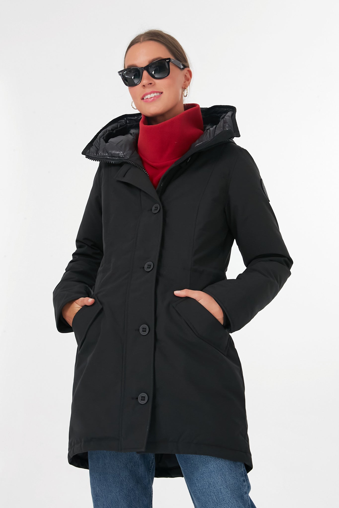A person in a black Canada Goose parka, red turtleneck, blue jeans, sunglasses, and pearl earrings stands confidently with hands in coat pockets.