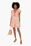 A woman in a Poupette St Barth Monaco Camila Mini Dress—floral, ruffled, boho-chic with pink trim—holds a woven bag and wears sandals for effortless French Riviera-inspired style.