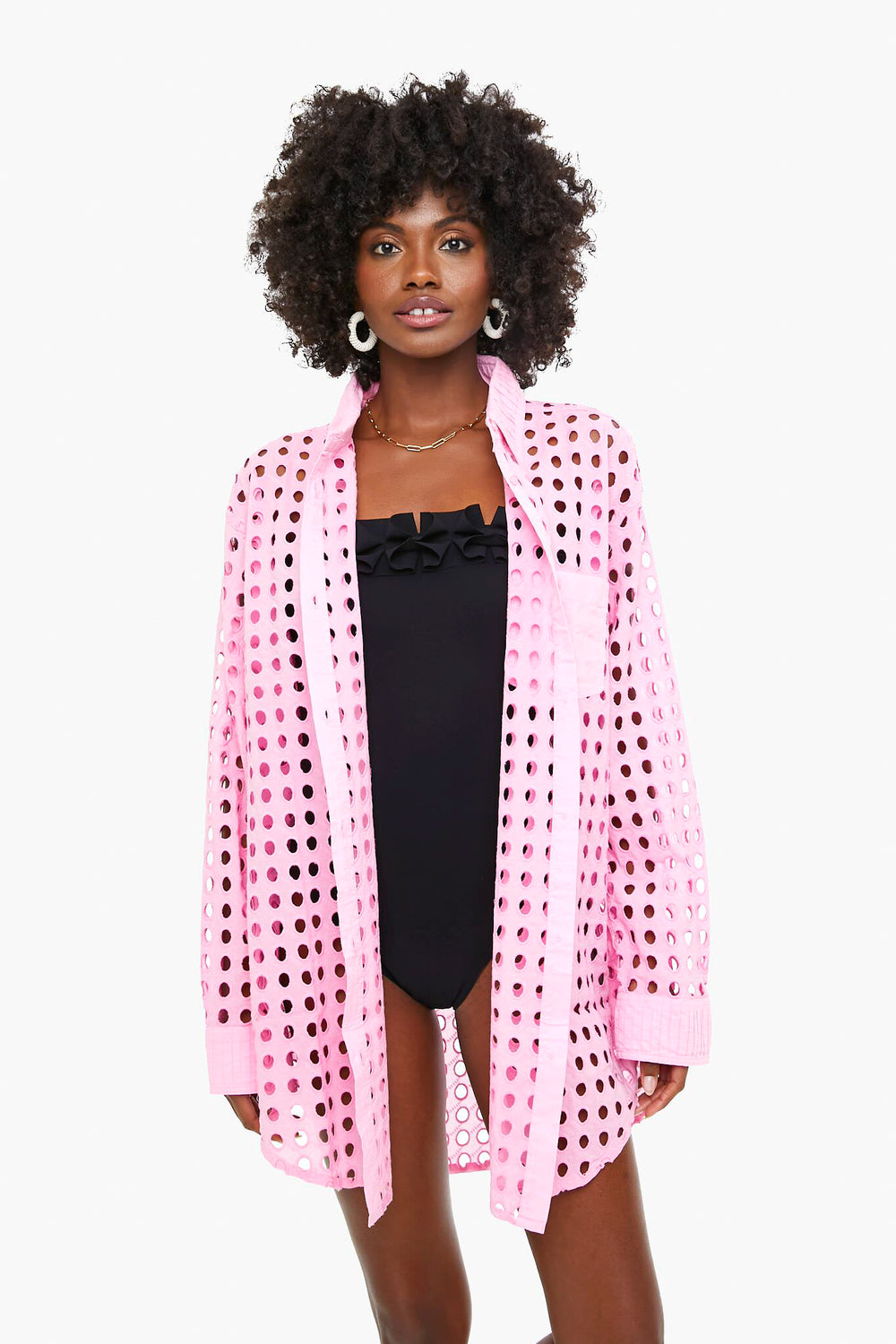 A woman wears a Solid & Striped black one-piece swimsuit with an oversized pink perforated button-up shirt styled as a beach coverup, standing facing forward.