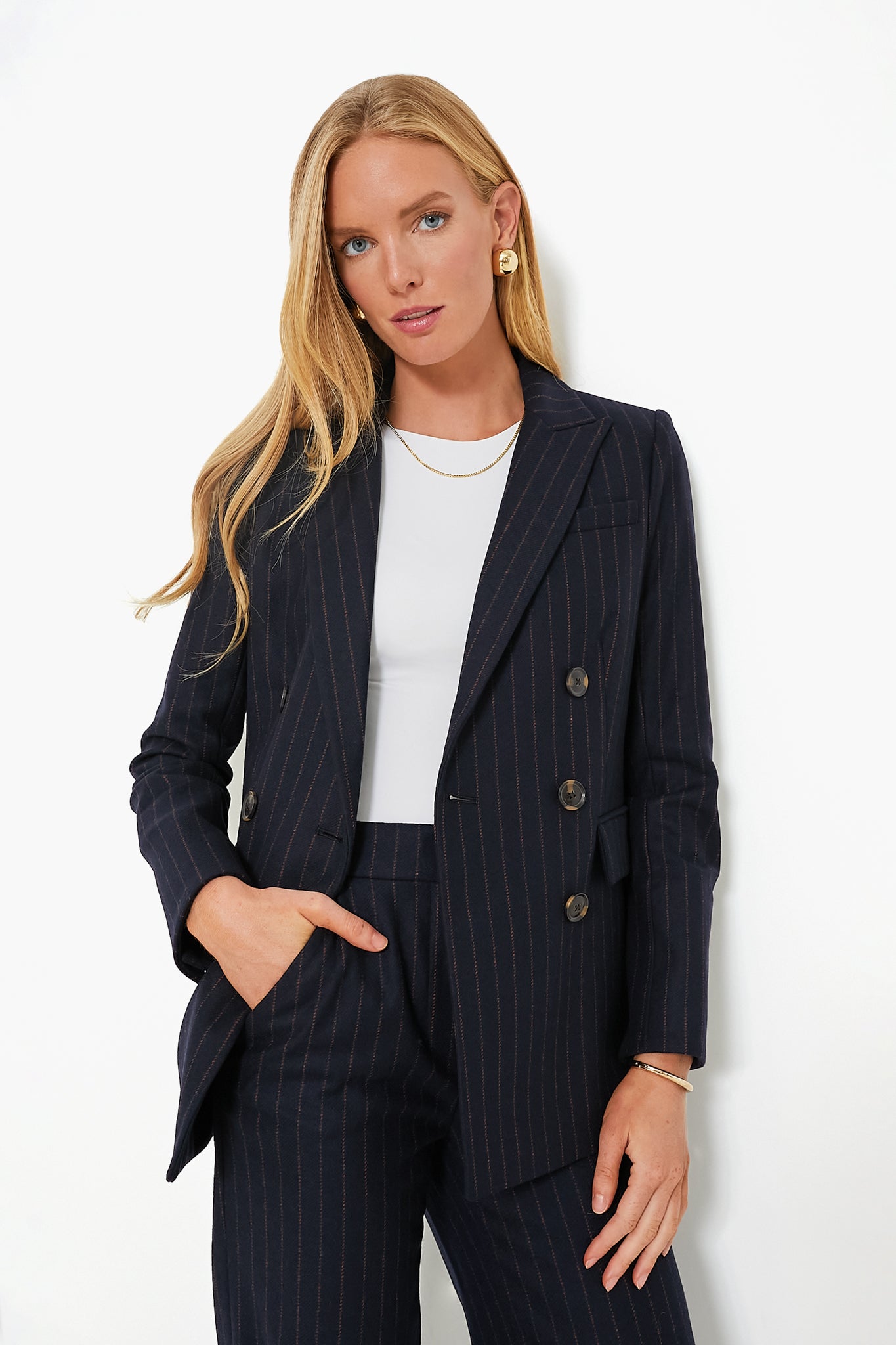 A woman stands confidently with one hand in her pocket, wearing a Veronica Beard navy pinstripe double-breasted blazer over a white top, accessorized with gold earrings and a necklace.