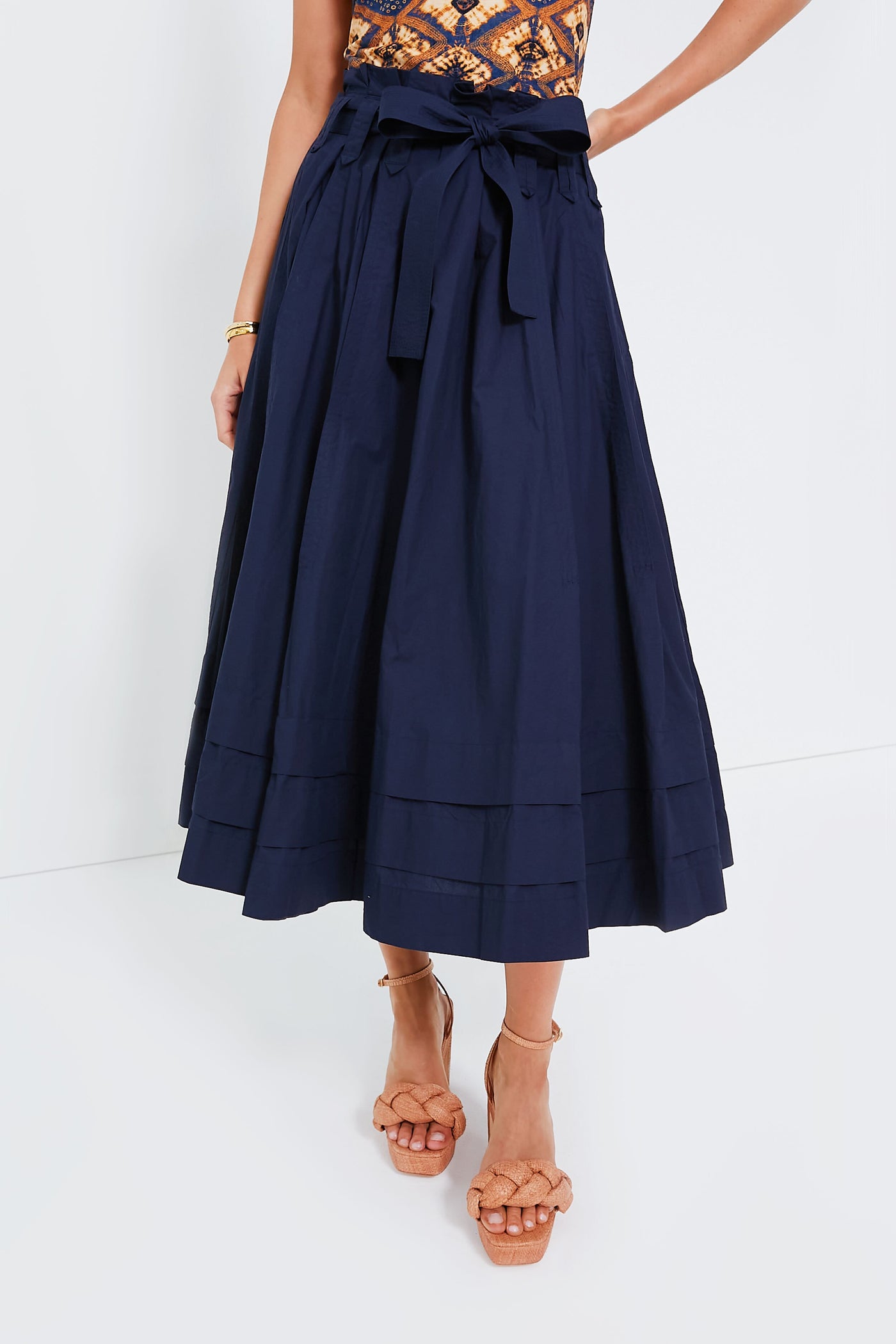 A woman wears a patterned sleeveless top, a Ulla Johnson navy blue cotton poplin paper bag waist midi skirt with a bow tie, and tan braided platform sandals.