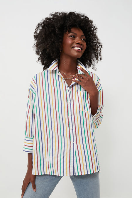 A woman wears a Pomander Place rainbow striped women’s button down top and light blue jeans, smiling and looking slightly to the side.