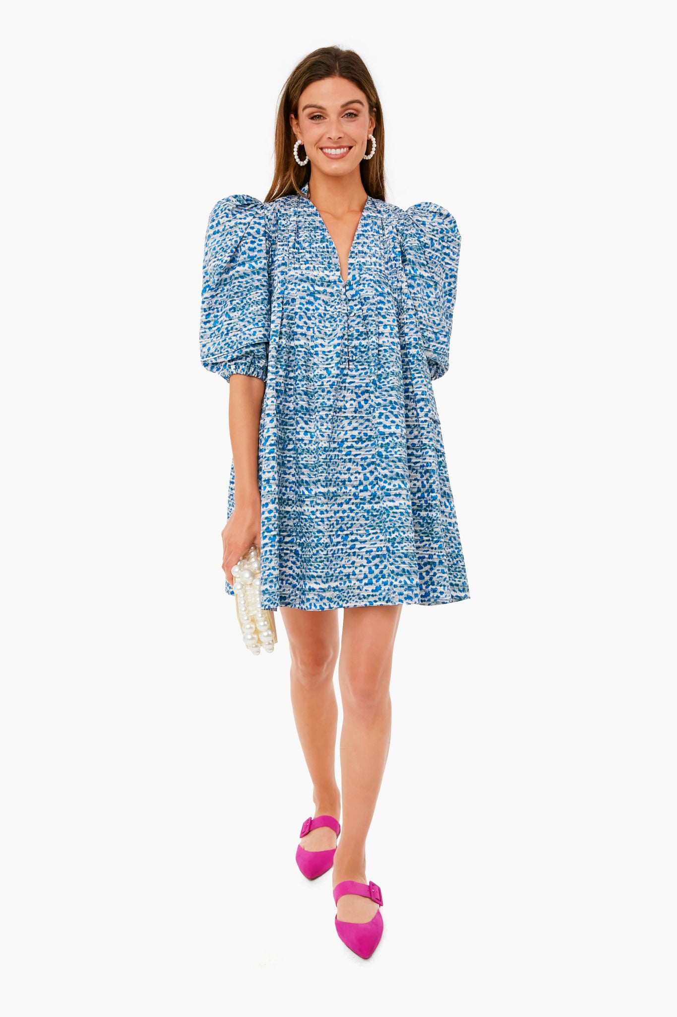 A woman wears a Hunter Bell Jenkins Dress featuring dramatic puff shoulders in blue and white leopard print, styled with pink shoes and a white beaded clutch.