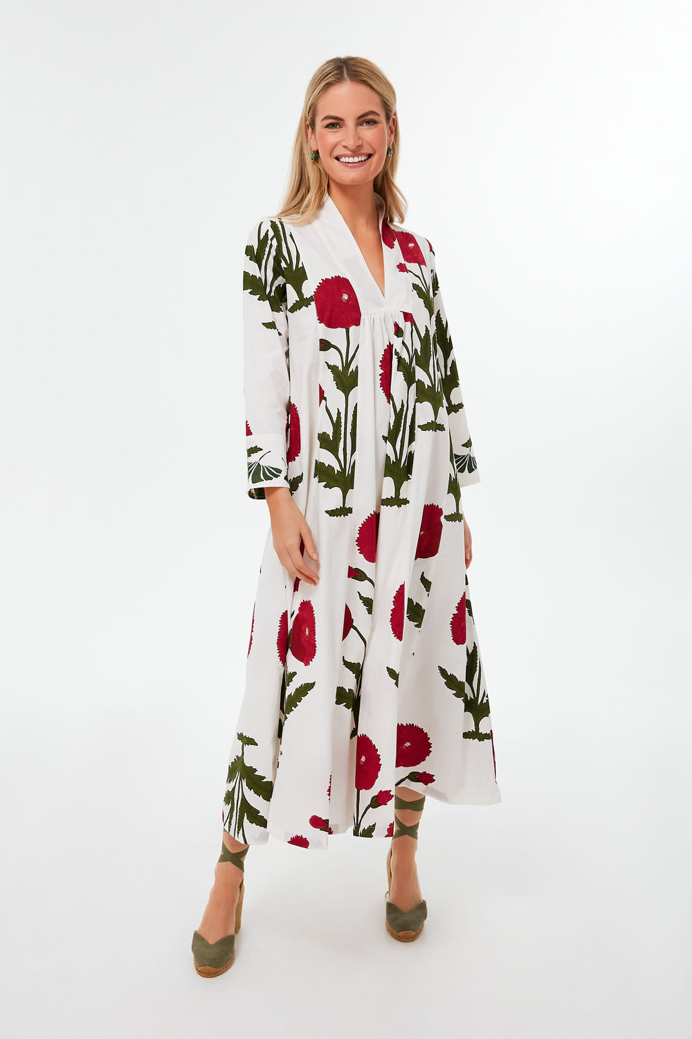 A woman smiles in a long white cotton KLEED kaftan with a red and green floral print, paired with green heels.