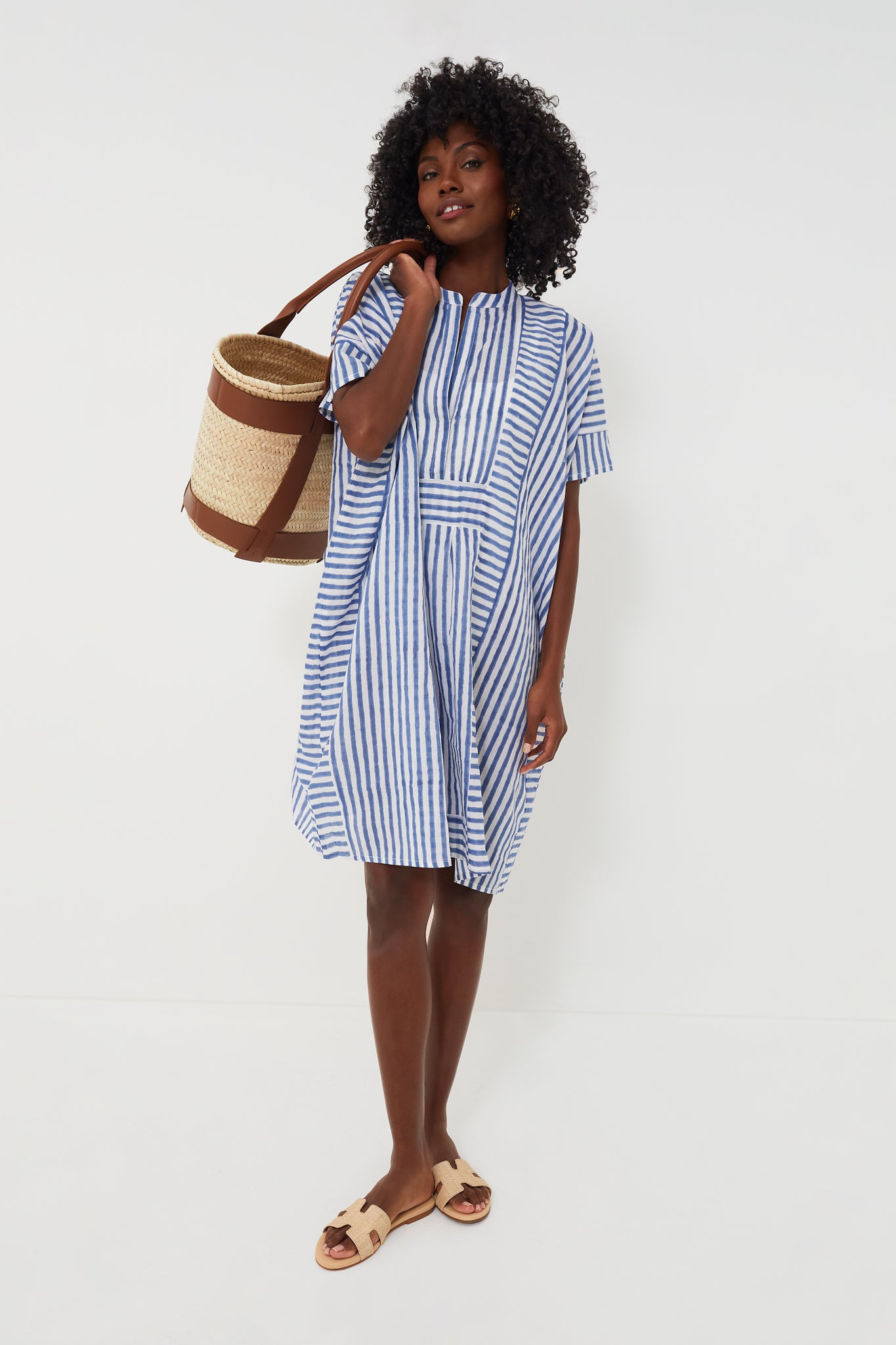A woman stands wearing a blue and white striped short-sleeve Emerson Caftan in organic cotton by Love Tomas by Emerson Fry, paired with tan sandals and carrying a straw tote bag.