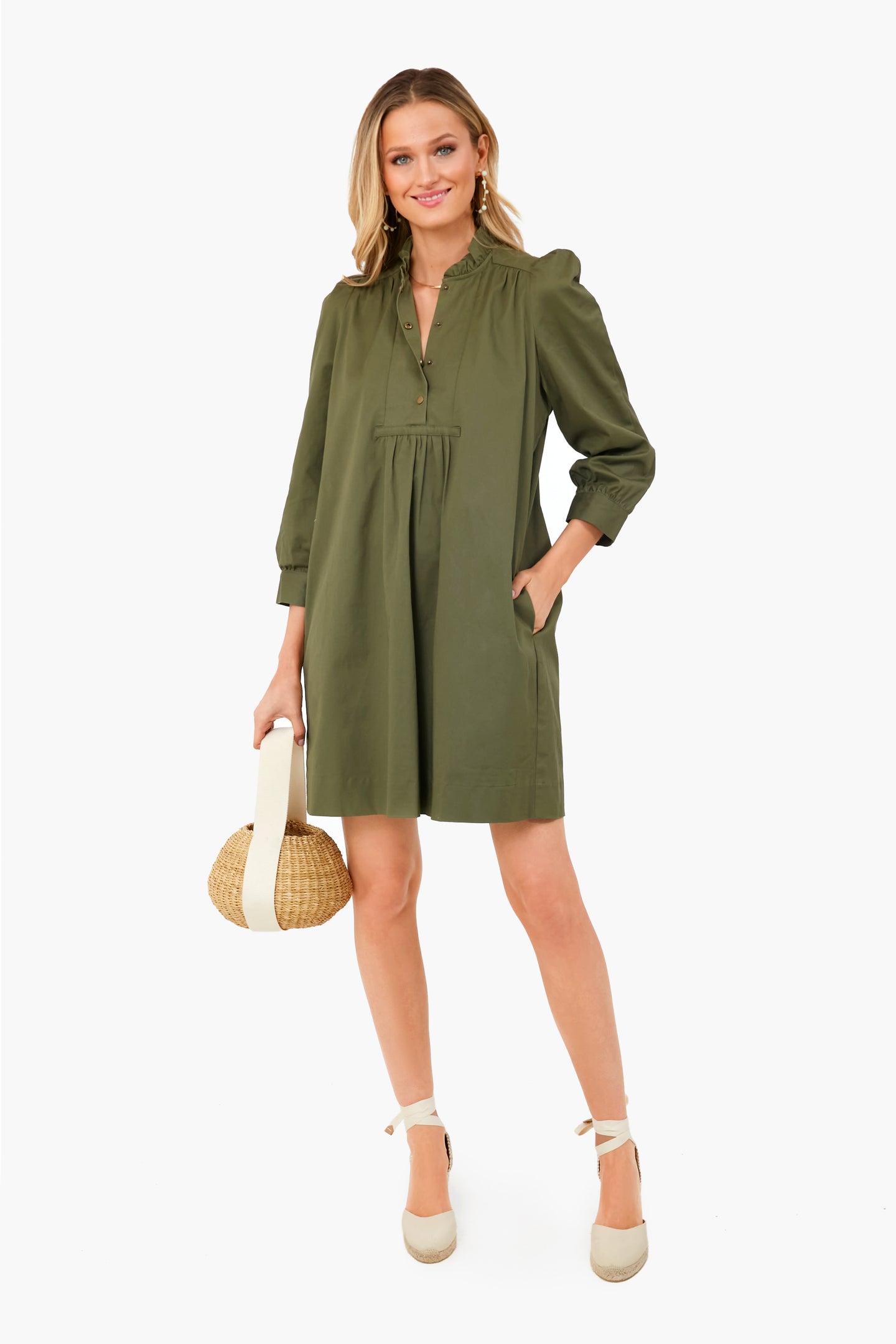 A woman wears an olive green long-sleeve Pomander Place dress with puff sleeves and pockets, paired with a round straw handbag and beige espadrille wedges.
