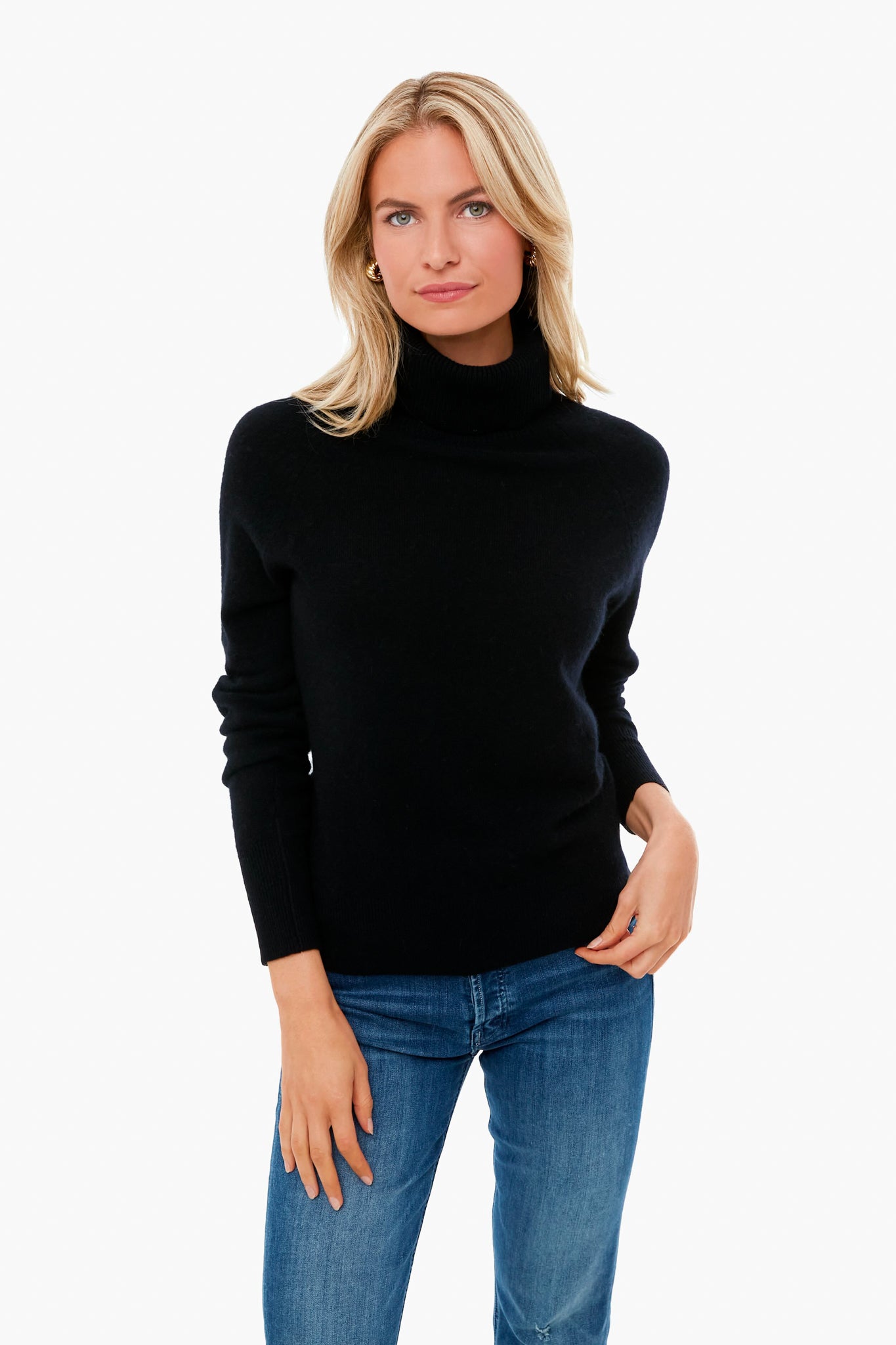 A blonde woman wears a White + Warren black cashmere turtleneck sweater with blue jeans, standing in a minimalist setting.
