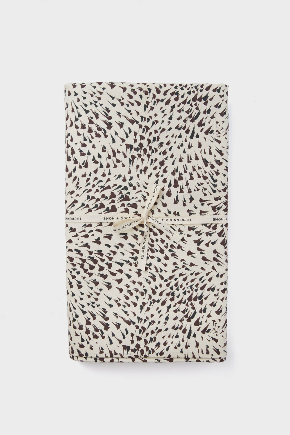A folded cotton tablecloth with a black and white spotted pattern, tied with a cream ribbon printed with "Tuckernuck Home." Stylish and practical, this machine washable tablecloth adds charm to any setting.