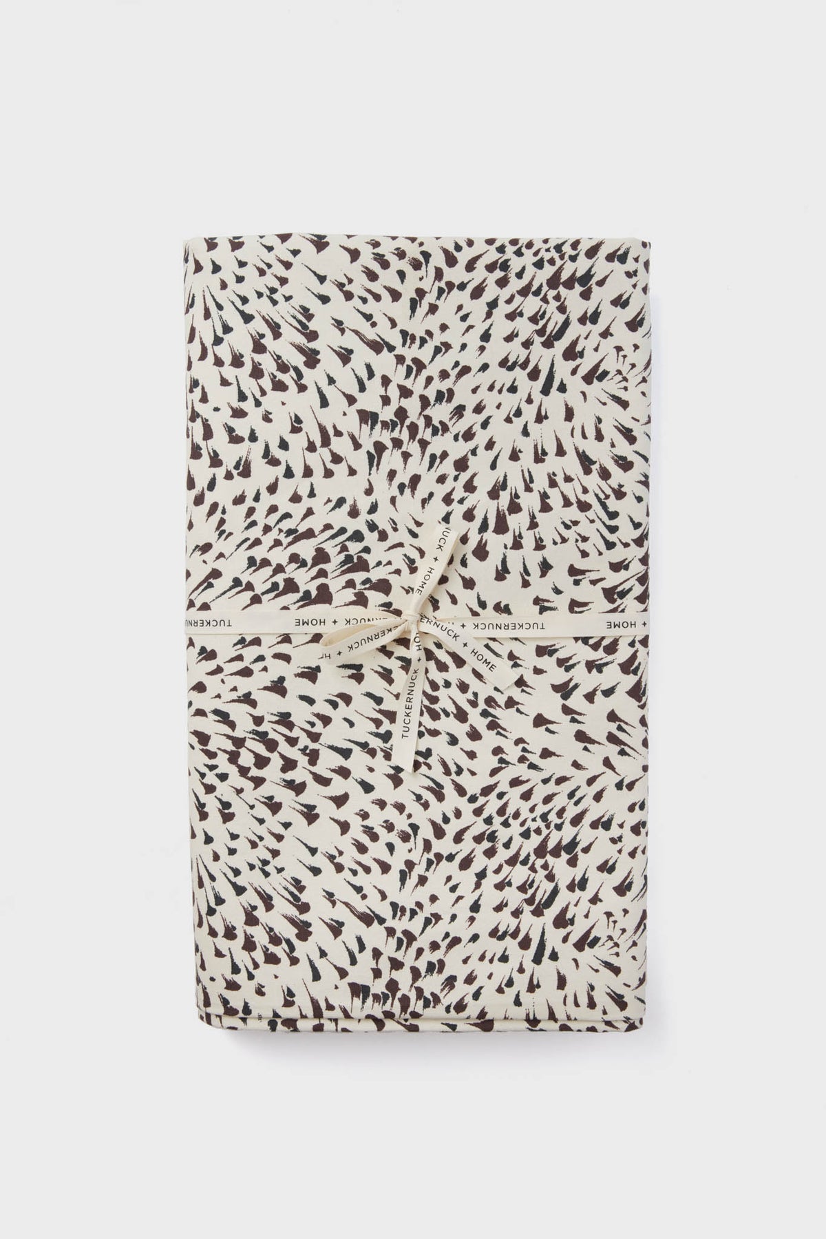 A folded cotton tablecloth with a black and white spotted pattern, tied with a cream ribbon printed with "Tuckernuck Home." Stylish and practical, this machine washable tablecloth adds charm to any setting.