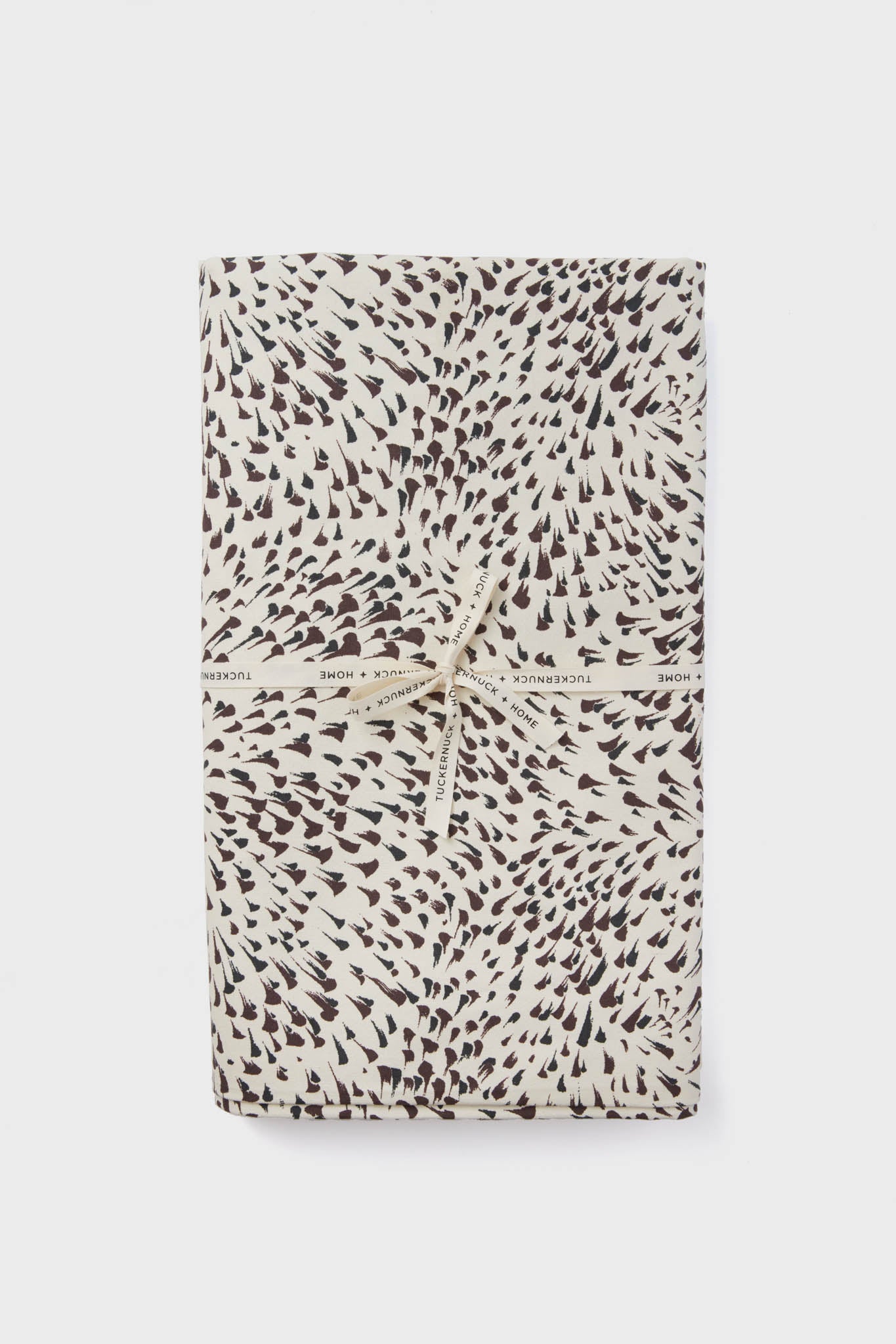 A folded cotton tablecloth with a black and white spotted pattern, tied with a cream ribbon printed with "Tuckernuck Home." Stylish and practical, this machine washable tablecloth adds charm to any setting.