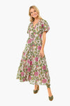 A smiling woman wears a Banjanan long, flowy boho-chic dress with a pink and green floral pattern and short sleeves, crafted from organic cotton voile for comfort and style.