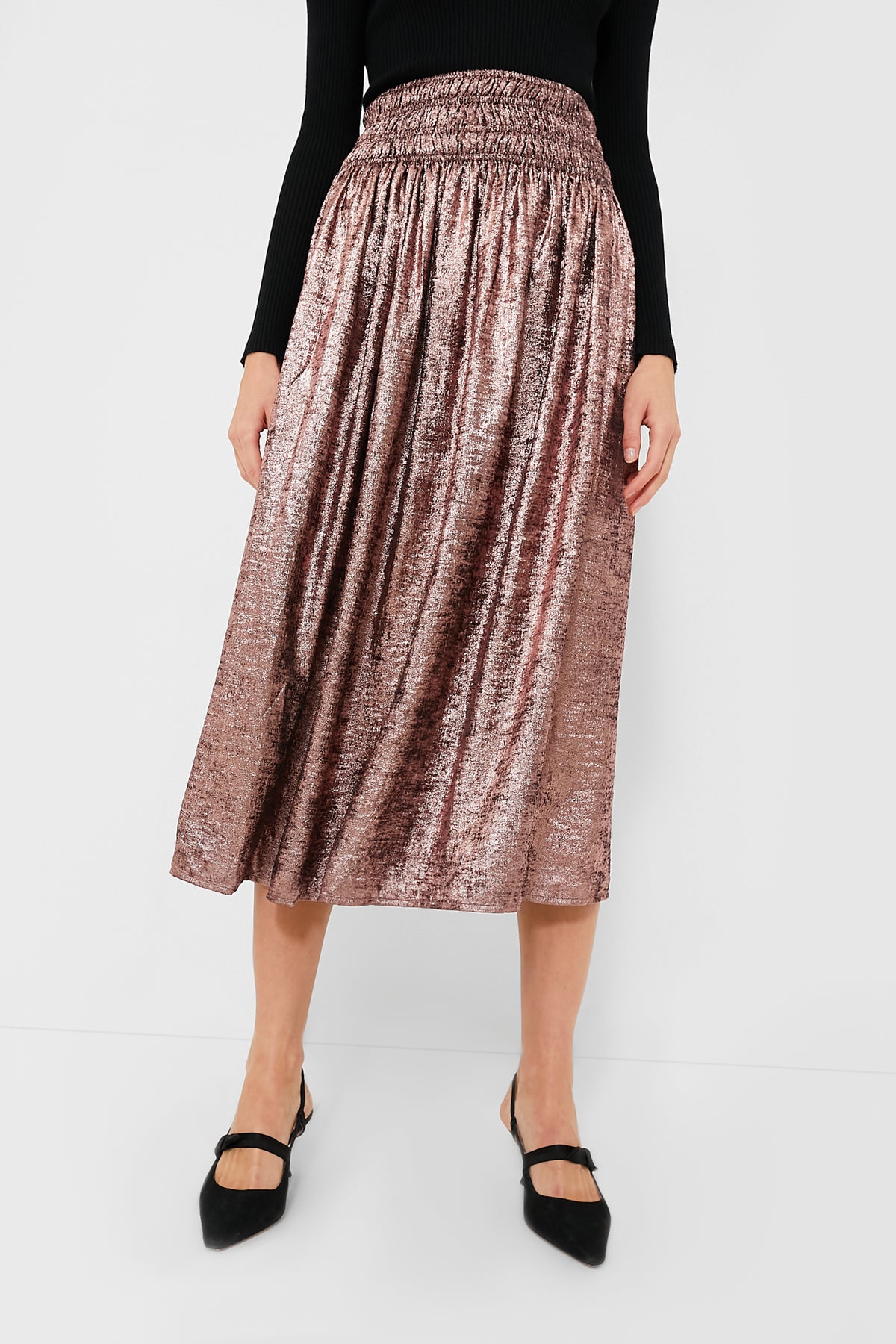 A person wears THE GREAT. shiny, flowy Rose Gold skirt with a black long-sleeve top and black pointed-toe shoes.