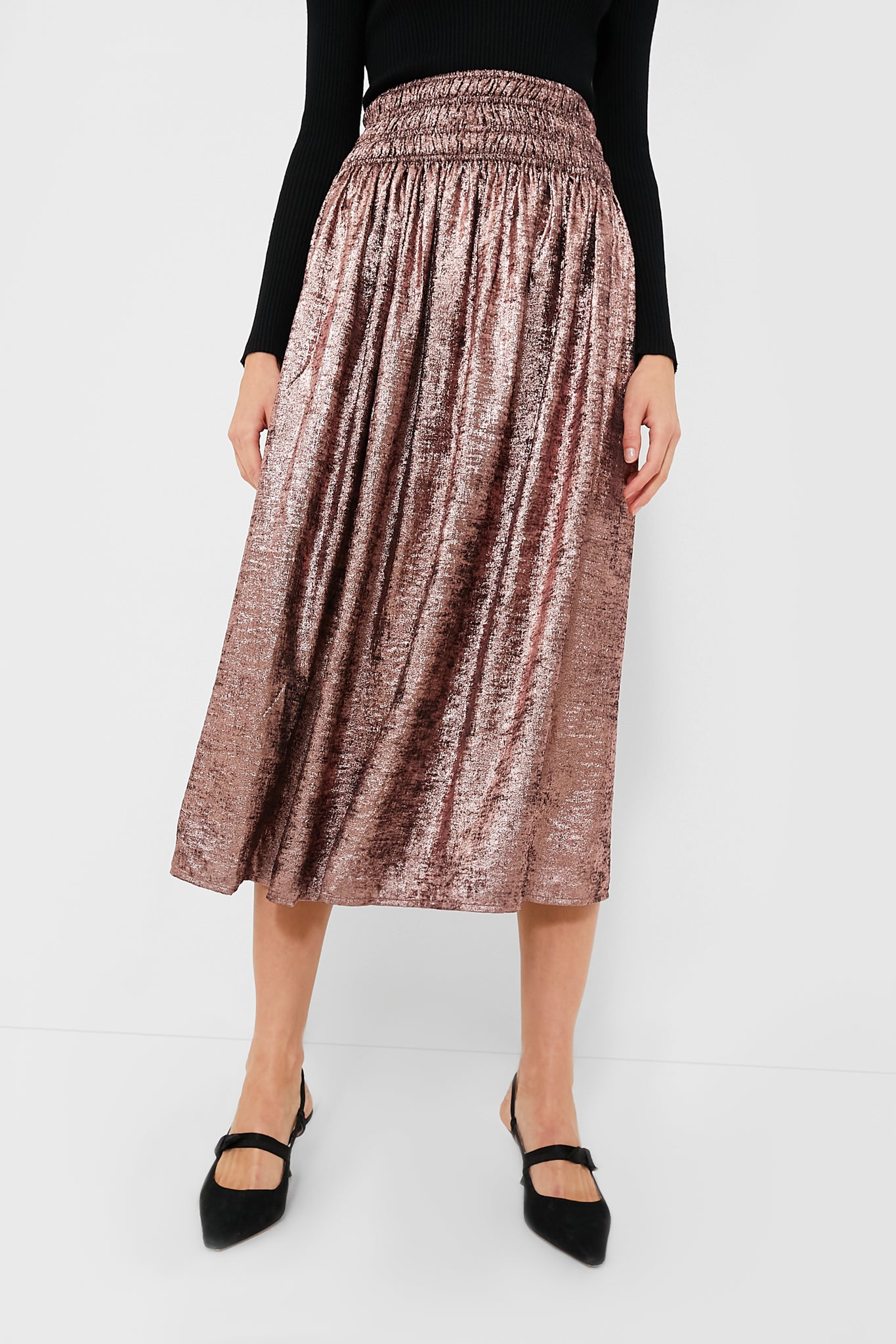 A person wears THE GREAT. shiny, flowy Rose Gold skirt with a black long-sleeve top and black pointed-toe shoes.