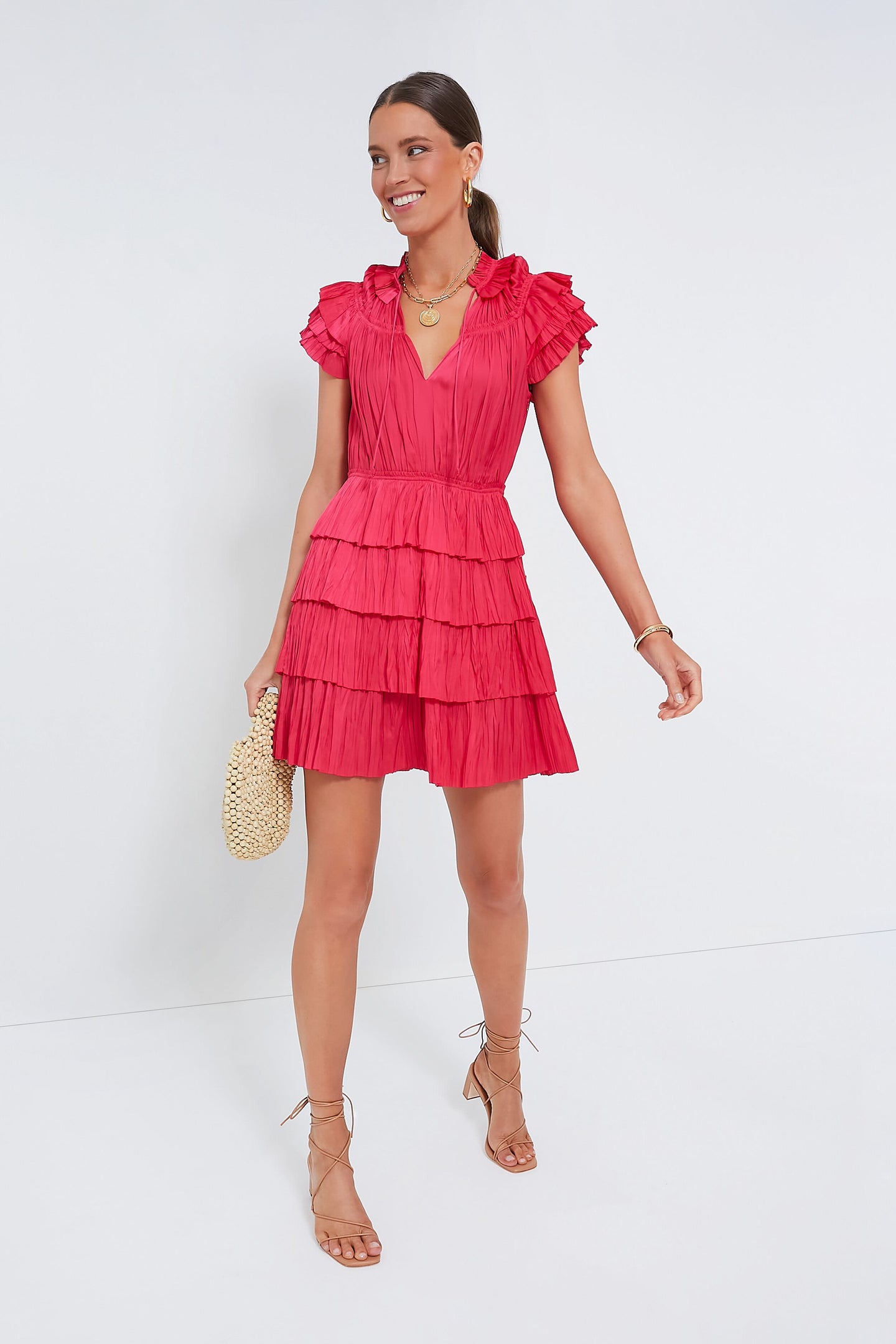 A woman stands poised and stylish in a Ulla Johnson short, tiered red ruffle mini dress with cap sleeves, holding a small woven bag and wearing sandals.