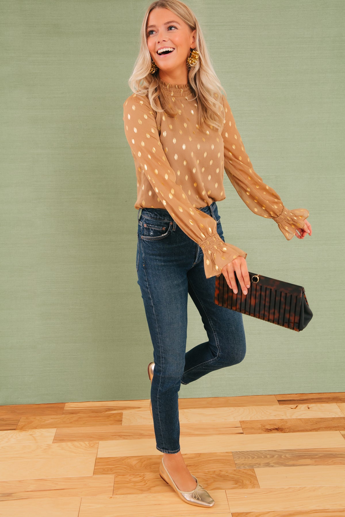 Model mid-stride, wearing classic metallic gold ballet flats and holding the high-end Tortoise Iris Clutch, effortlessly pairing with fun, sheer blouse and dark jeans.