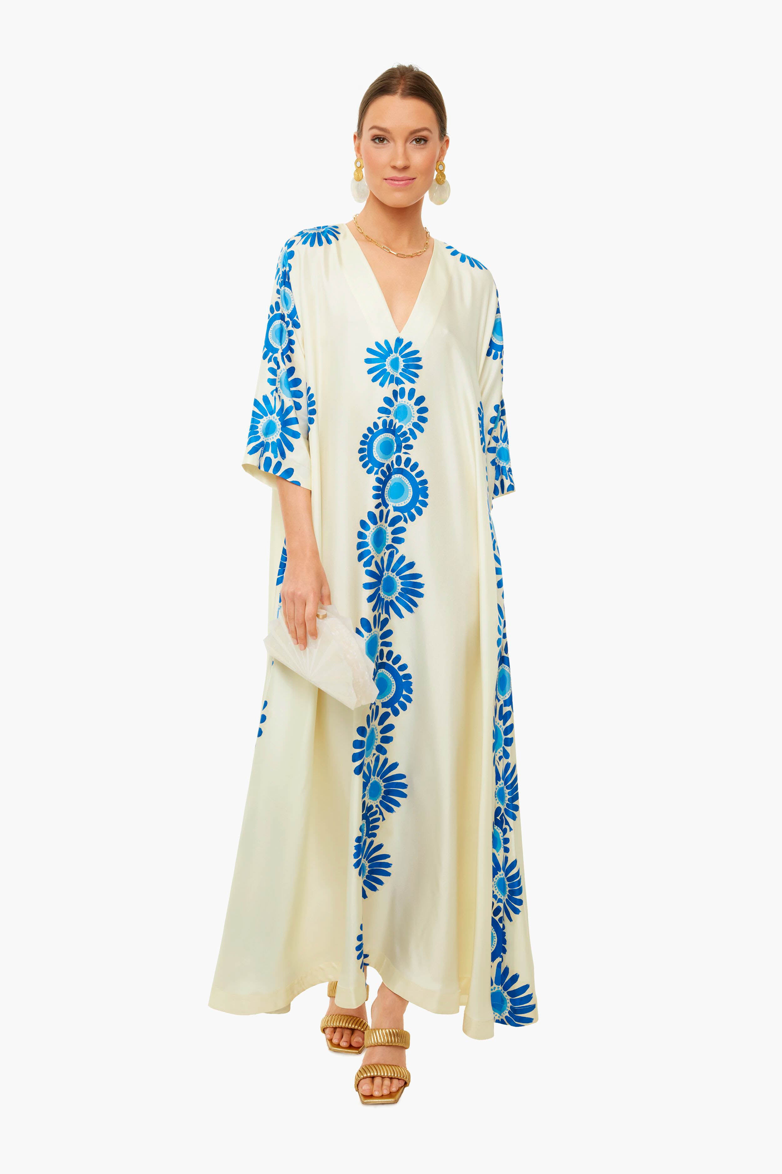 A woman wears a La DoubleJ silk cream maxi dress with hand-printed daisy motifs, paired with a white clutch, tan sandals, and gold earrings. Made in Italy.