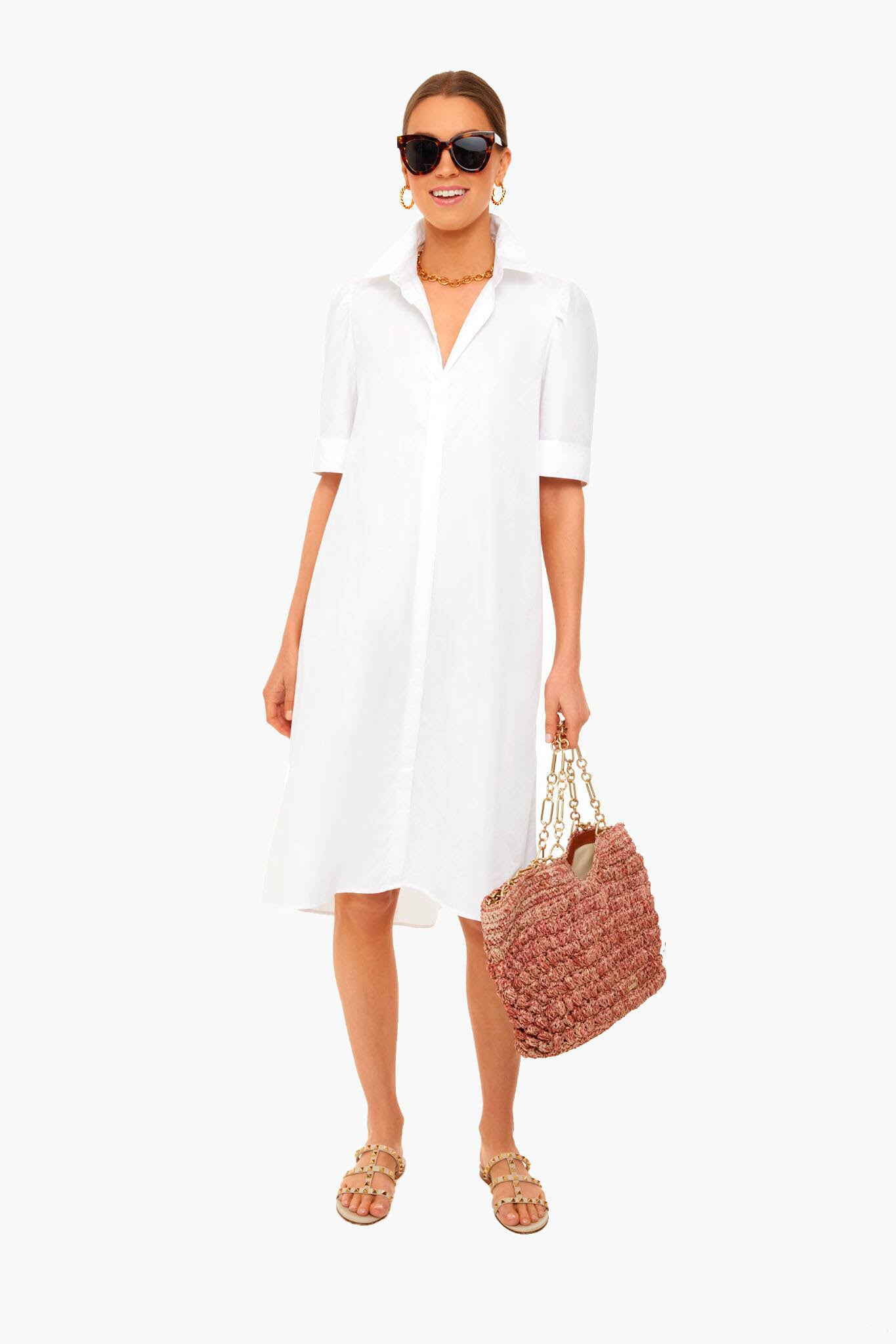 A woman in a Pomander Place white cotton linen shirtdress, sunglasses, gold earrings, necklace, and sandals carries a large woven handbag.