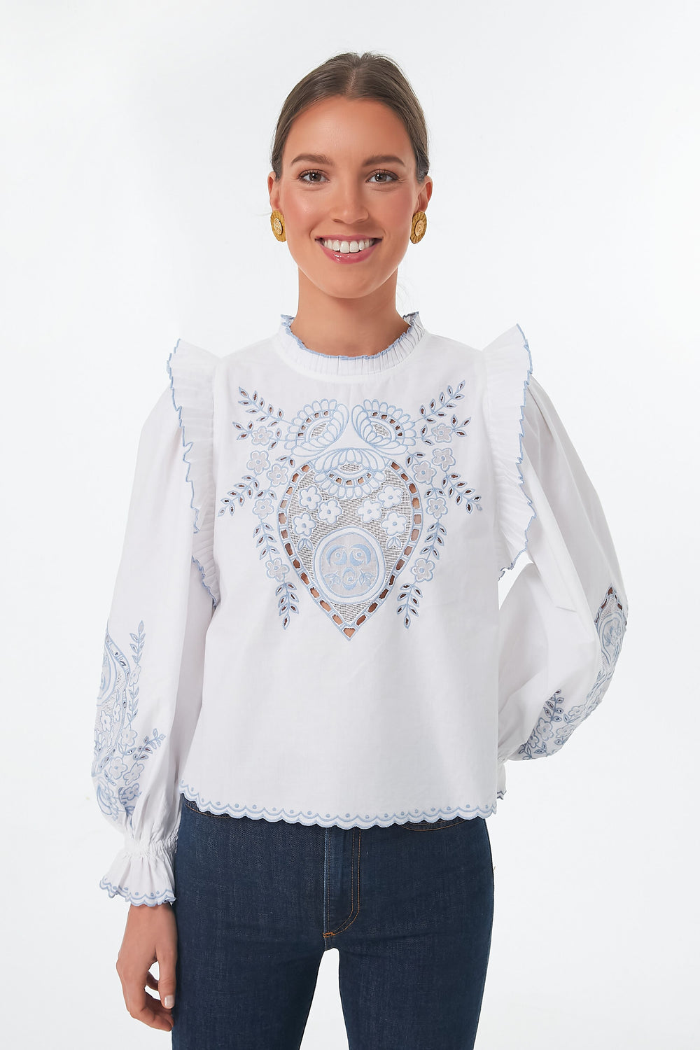 A woman wears a Love the Label cotton poplin blouse with blue embroidery, ruffled sleeves, and a pleated mock neck, paired with dark blue jeans.