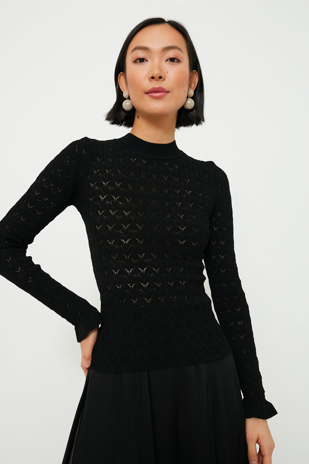 A person stands facing the camera wearing a black sheer knit long sleeve top and black skirt by Sea New York.