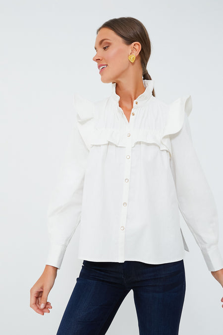 A woman wearing the Tuckernuck Winnie Blouse—a white cotton ruffle blouse with long sleeves—paired with blue jeans, looks to her left.