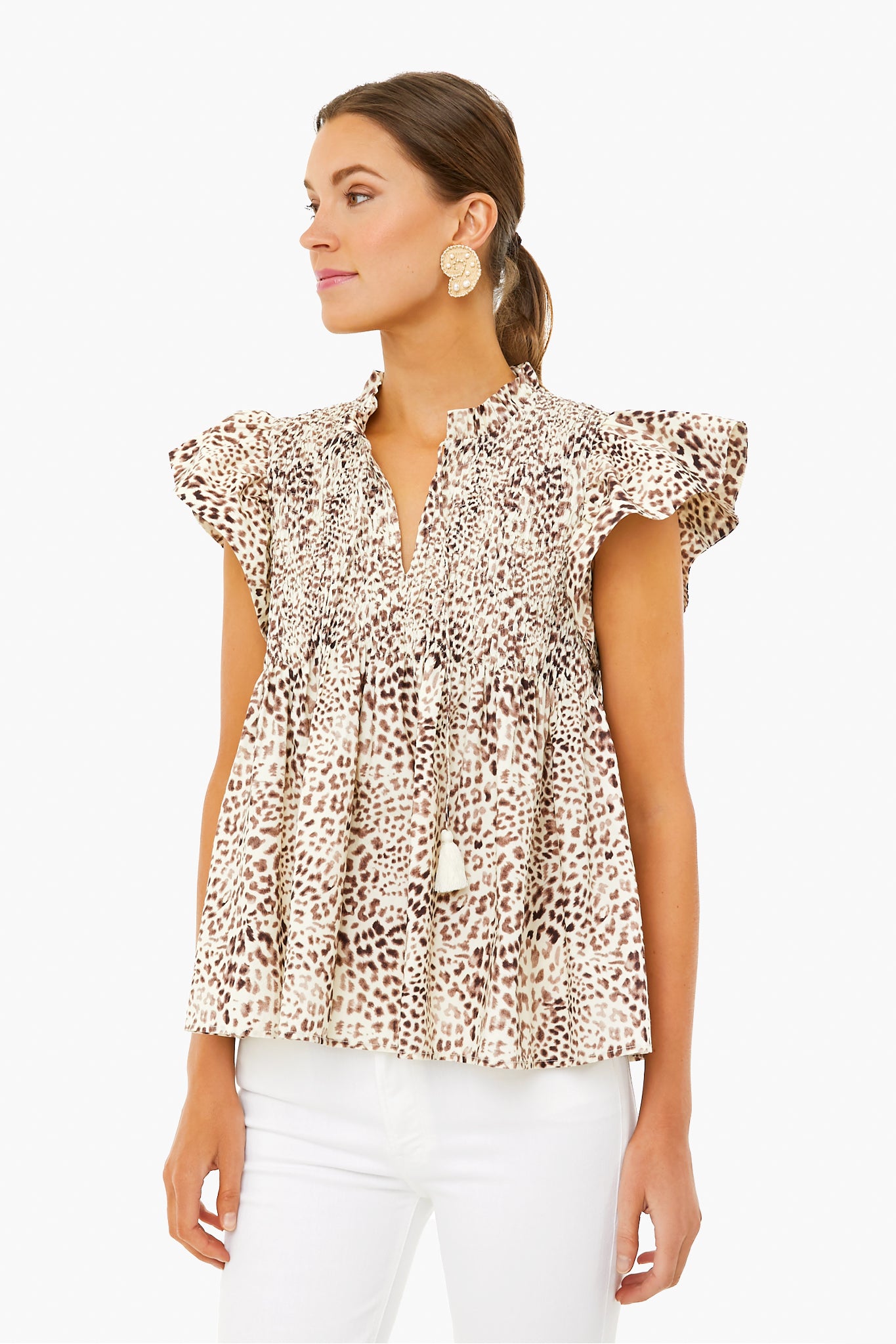 Woman in a Sea New York cheetah-print smocked top with flutter sleeves, ruffles, and tassel ties—styled with white pants and round statement earrings for a modern look.