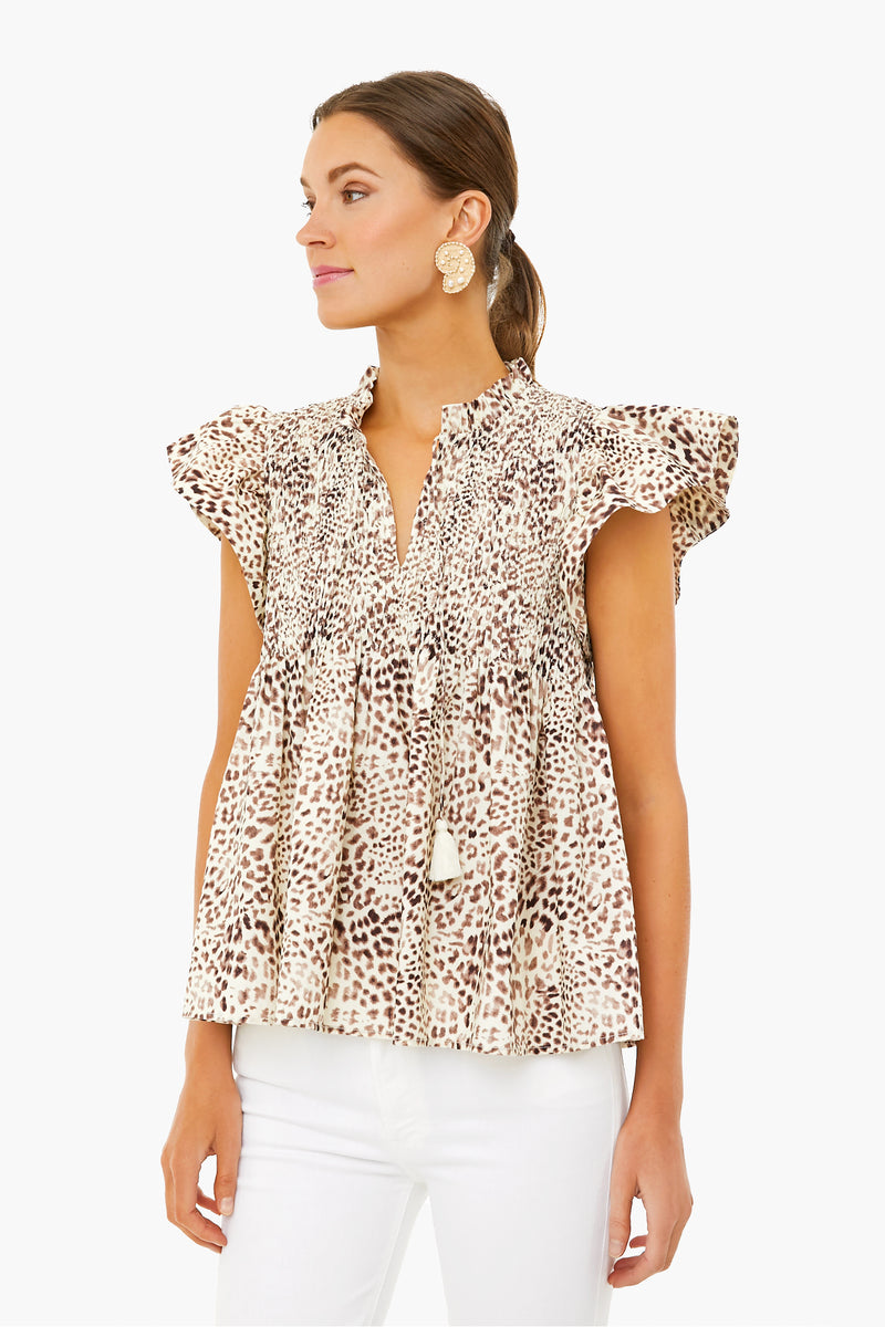 Woman in a Sea New York cheetah-print smocked top with flutter sleeves, ruffles, and tassel ties—styled with white pants and round statement earrings for a modern look.