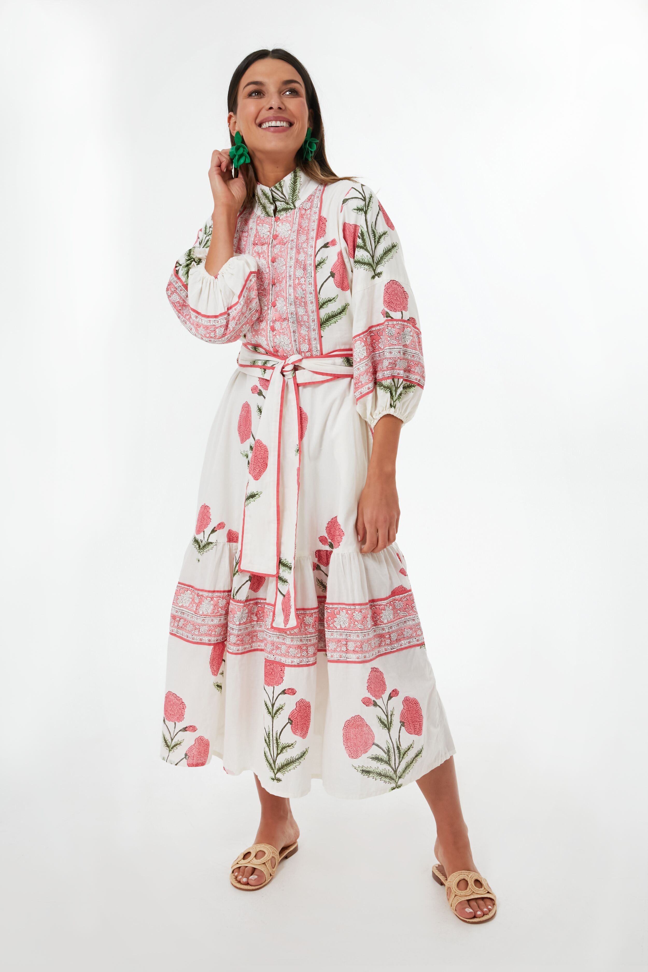 A woman smiles in a Sue Sartor Rosy Marigold Flounce Dress with pink florals, made from sustainable fabrics, paired with green earrings and beige slide sandals.