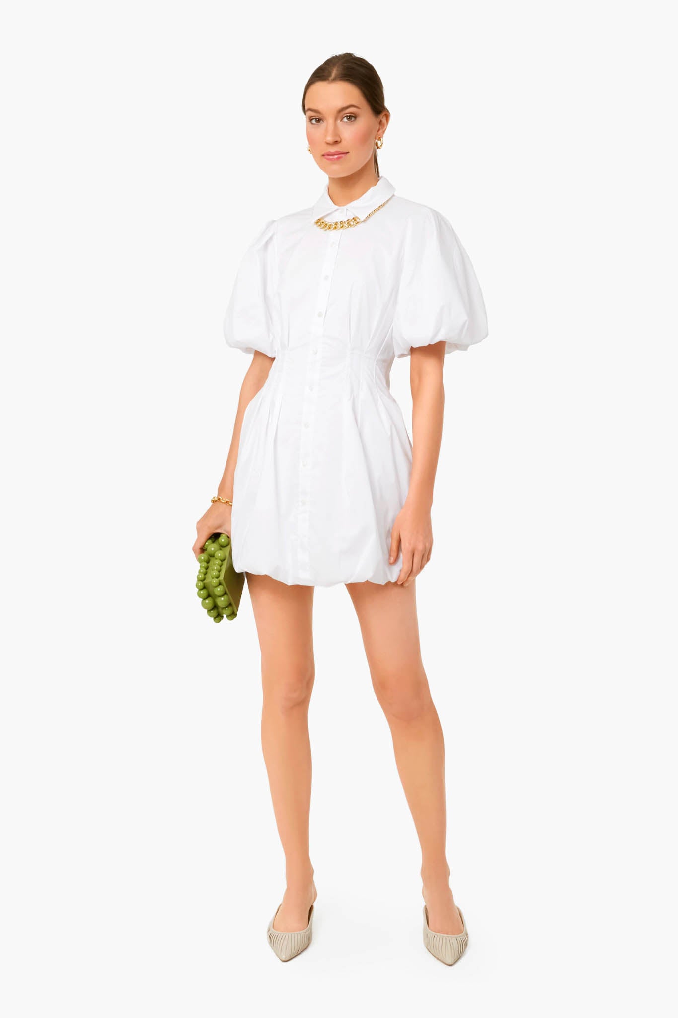 A woman in a SIMKHAI short white cotton poplin dress with puff sleeves, beige shoes, and gold jewelry holds green grapes, embodying luxury ready-to-wear style.