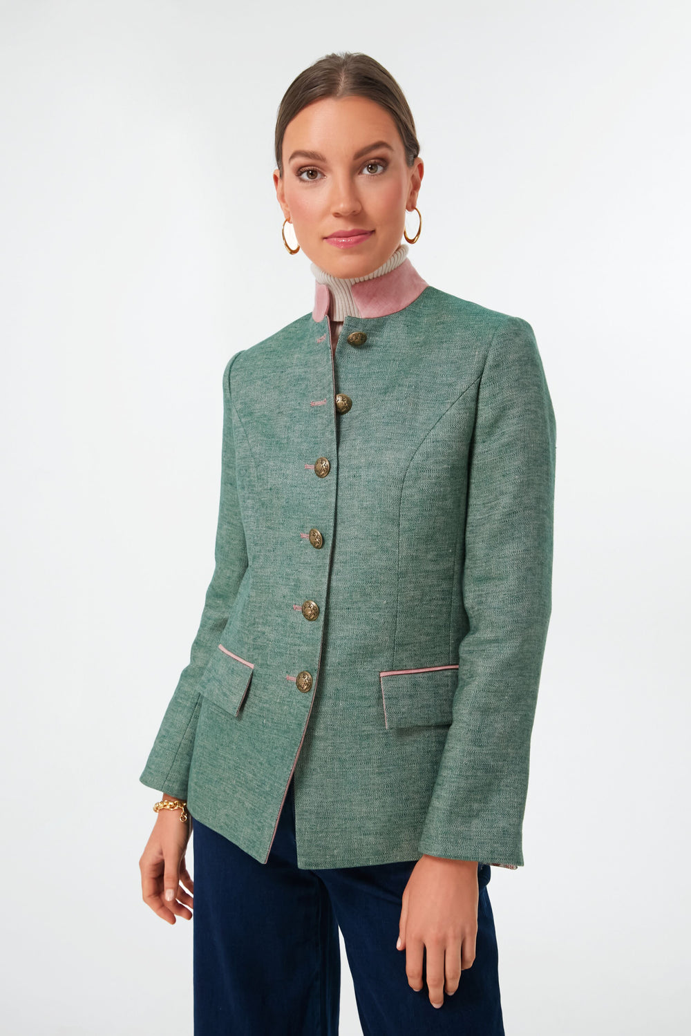 A woman wears a vintage-inspired green linen jacket with pink trim from Sanoe Collection, gold hoop earrings, and a pink high-neck top.