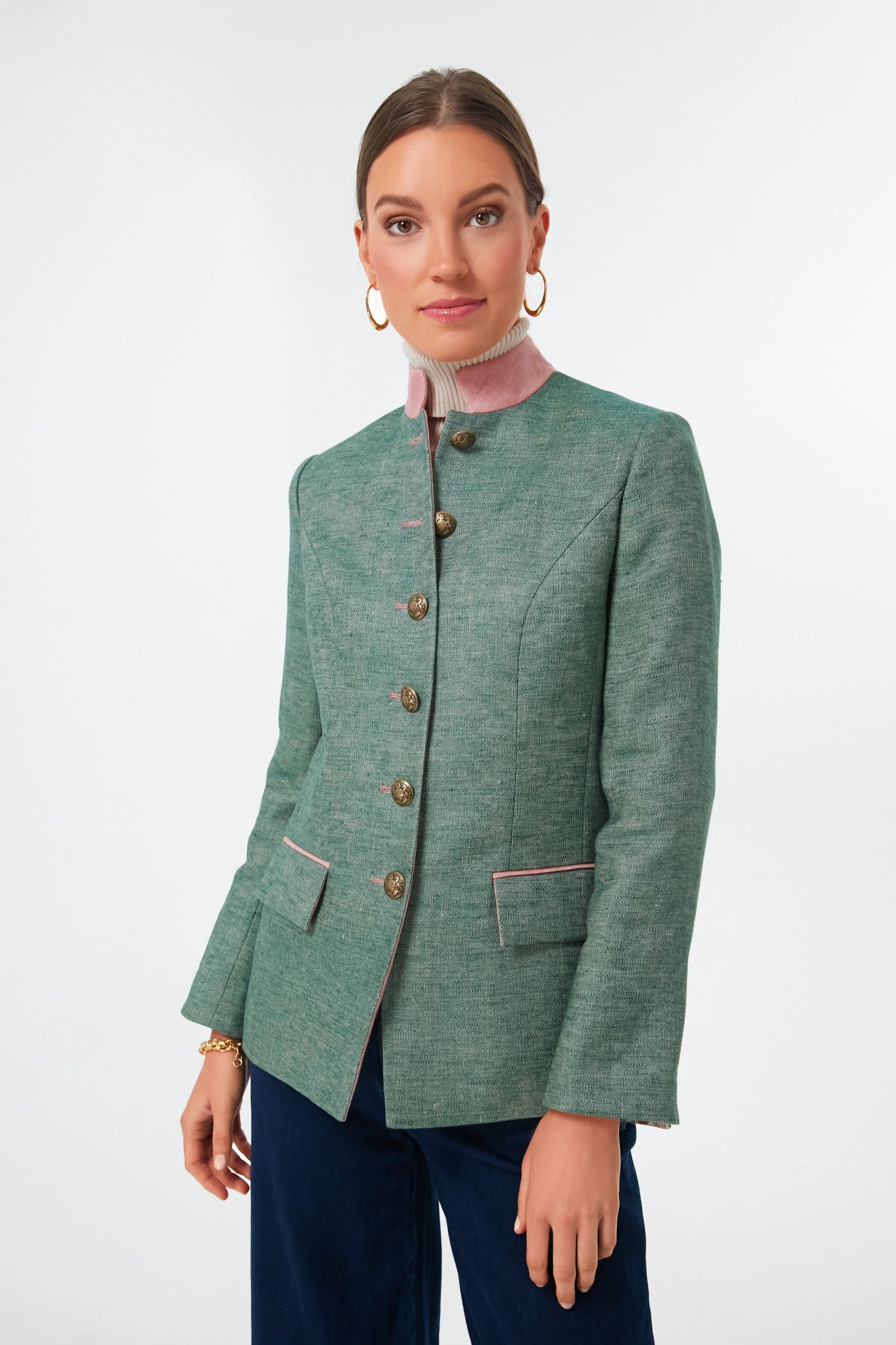 A woman wears a vintage-inspired green linen jacket with pink trim from Sanoe Collection, gold hoop earrings, and a pink high-neck top.