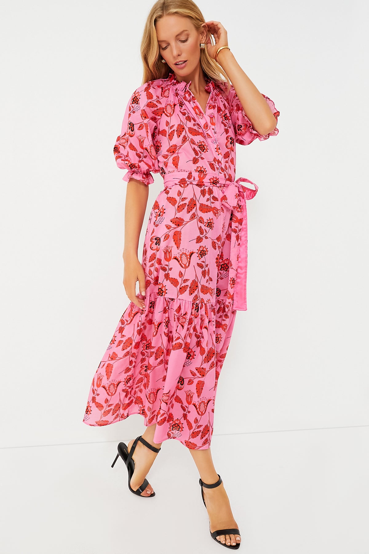 A woman poses in a Cynthia Rowley Saratoga Dress—an ankle-length pink wrap dress with red floral patterns, puffed sleeves, a tie waist, and black heeled sandals.