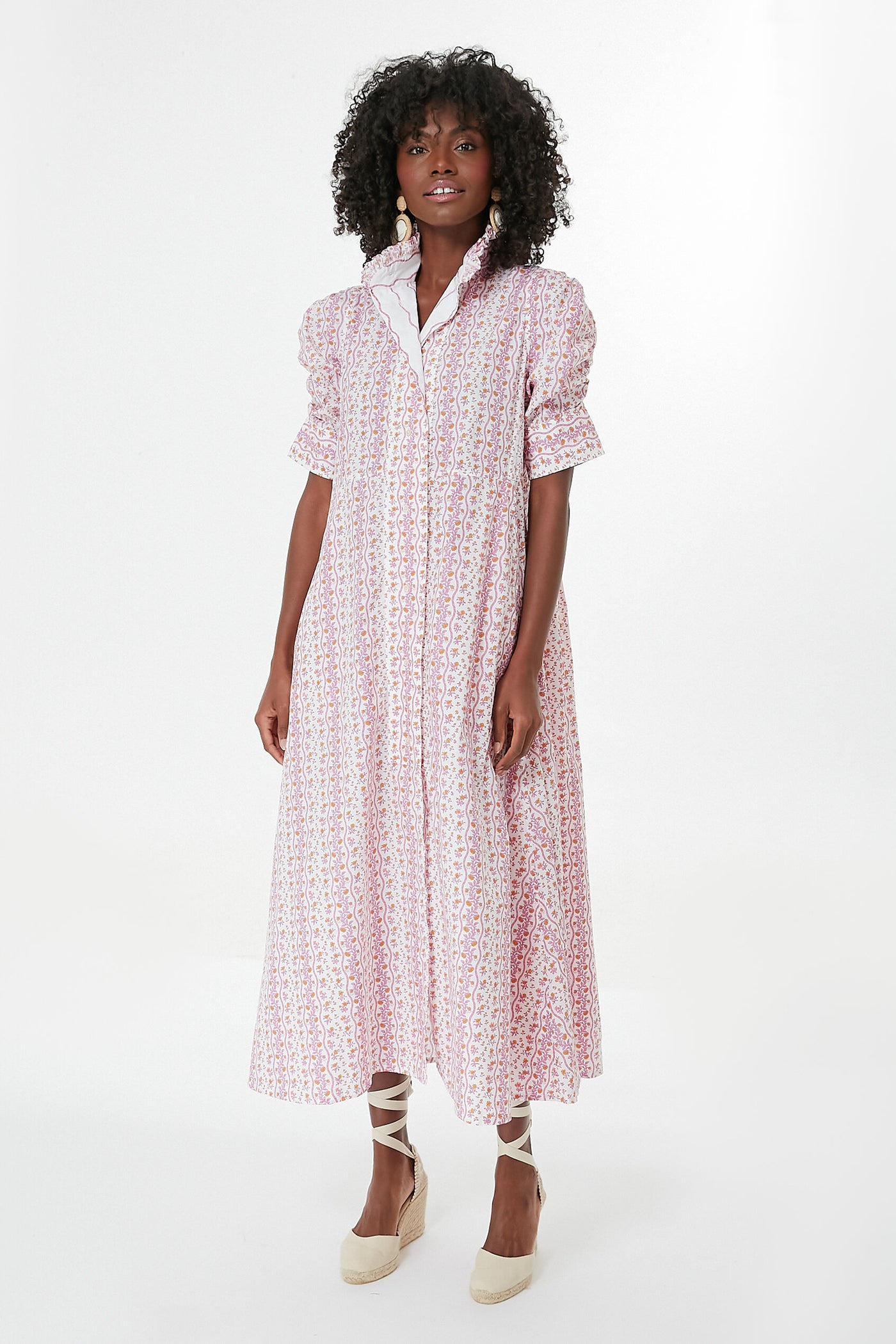 A woman wears a Thierry Colson pink floral midi dress with short puff sleeves and a high collar, paired with beige wedge espadrilles.