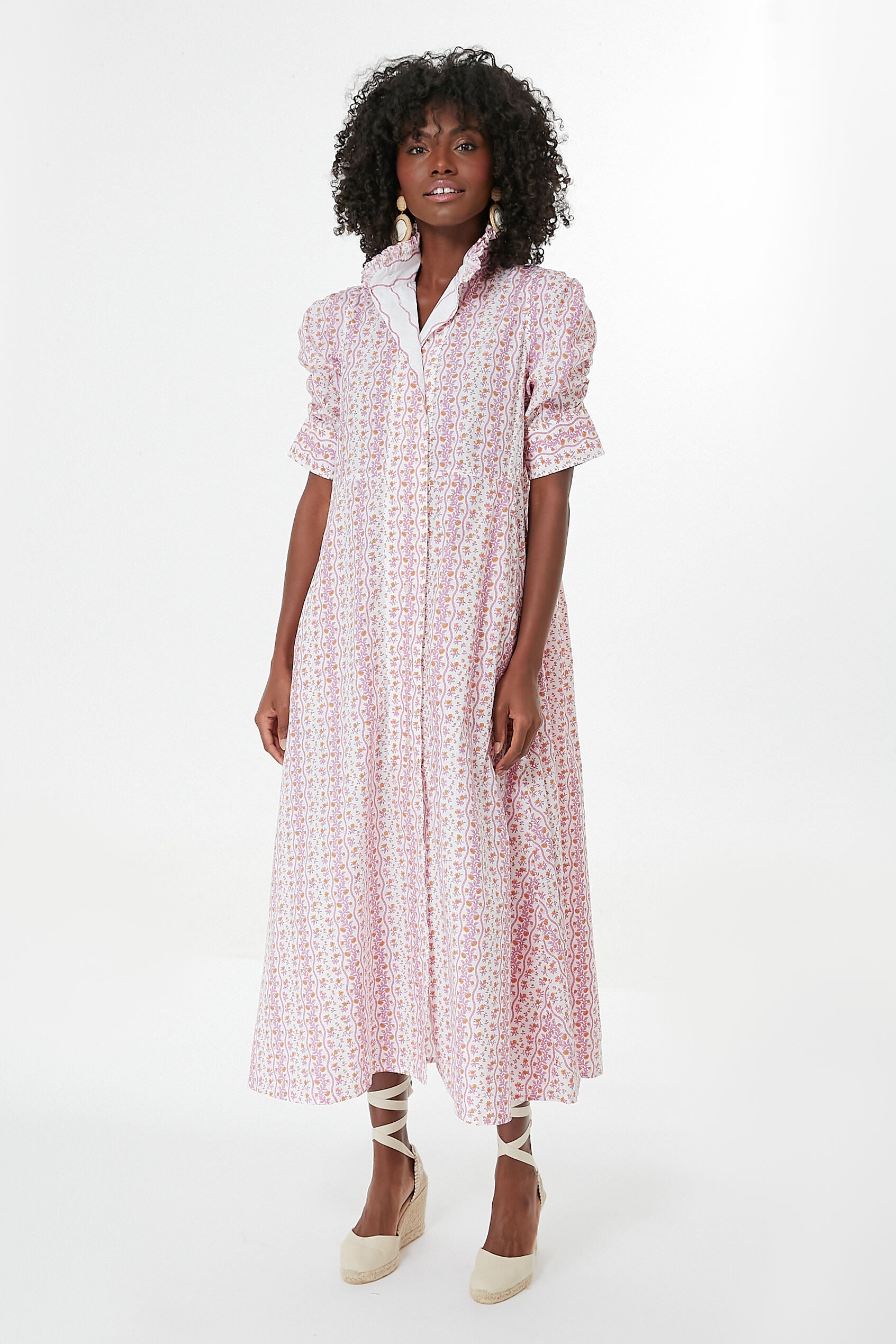 A woman wears a Thierry Colson pink floral midi dress with short puff sleeves and a high collar, paired with beige wedge espadrilles.