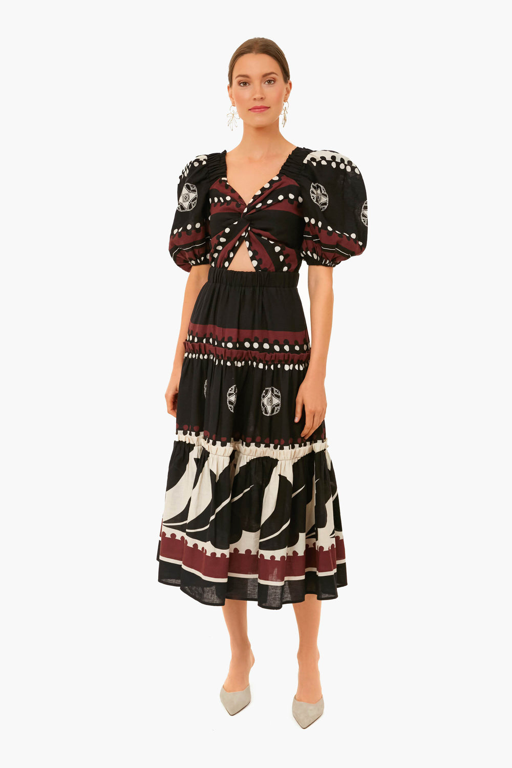 A woman wears the Black Liora Dress by Sea New York, a linen-cotton blend with white and burgundy patterns, puff sleeves, cut-out bodice, styled with beige pointed-toe shoes.