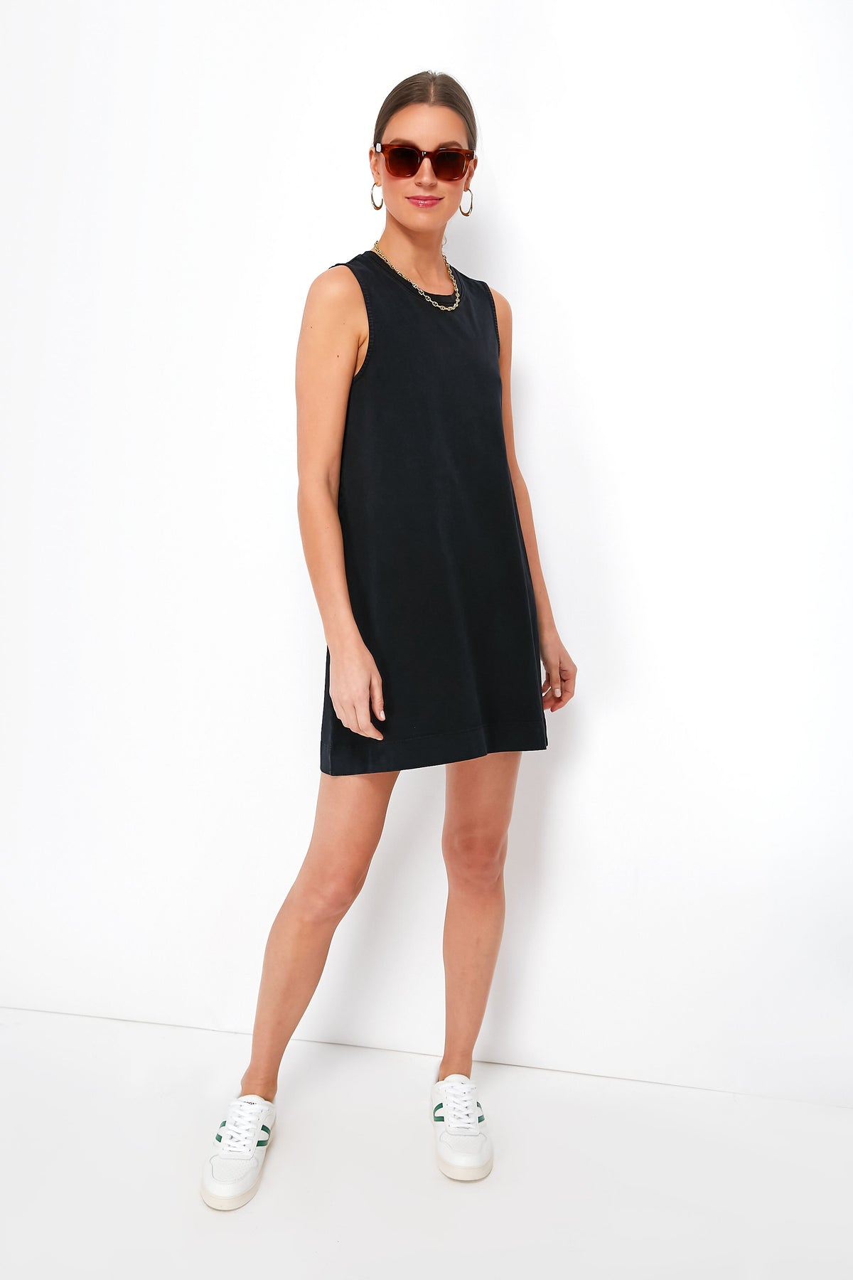 A woman in a sleeveless black Z SUPPLY Sloane dress, white sneakers, sunglasses, and hoop earrings stands confidently, showcasing effortless style.
