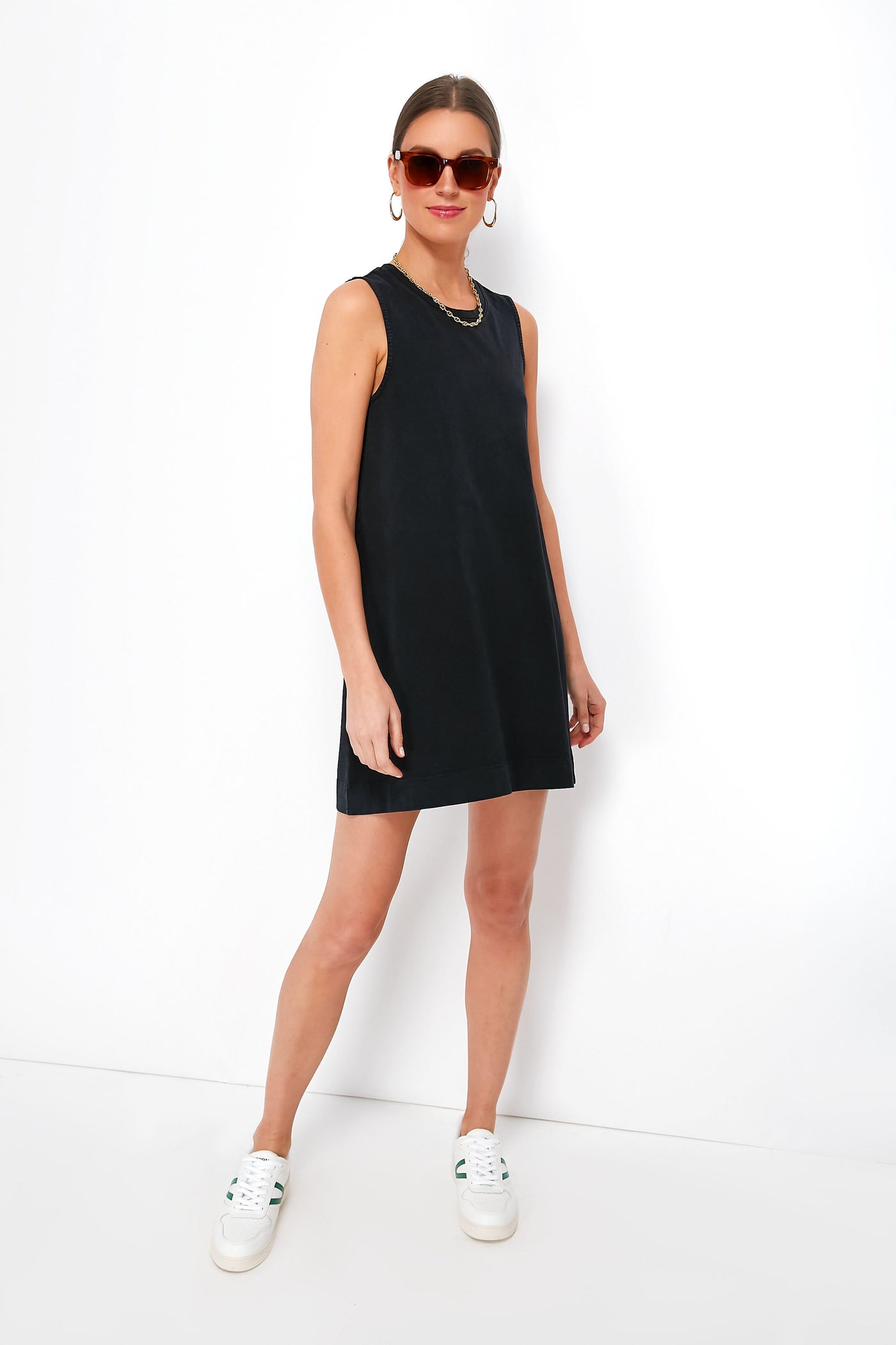 A woman in a sleeveless black Z SUPPLY Sloane dress, white sneakers, sunglasses, and hoop earrings stands confidently, showcasing effortless style.