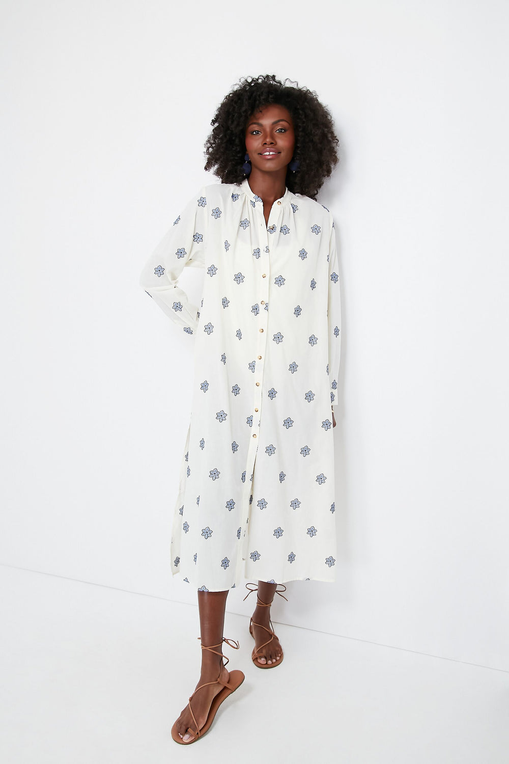A woman wears a long, white Ciao Lucia Gallo Dress with a blue floral print and brown strappy sandals, making it a chic beach cover-up.