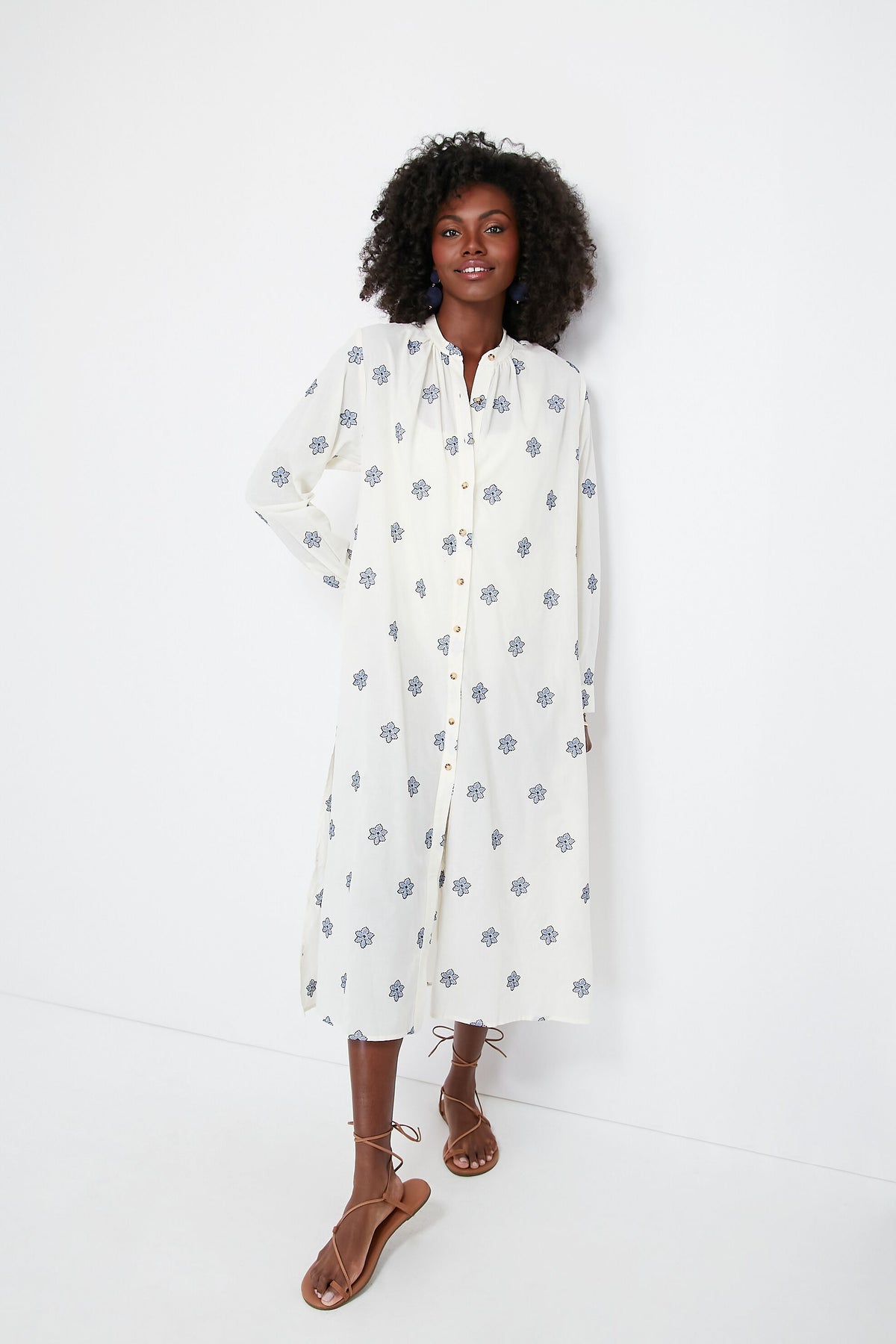 A woman wears a long, white Ciao Lucia Gallo Dress with a blue floral print and brown strappy sandals, making it a chic beach cover-up.