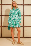 A woman indoors wears a vintage-inspired green chintz print long-sleeve dress with a pleated skirt from Tuckernuck, beige woven slip-on shoes, and holds a matching clutch.