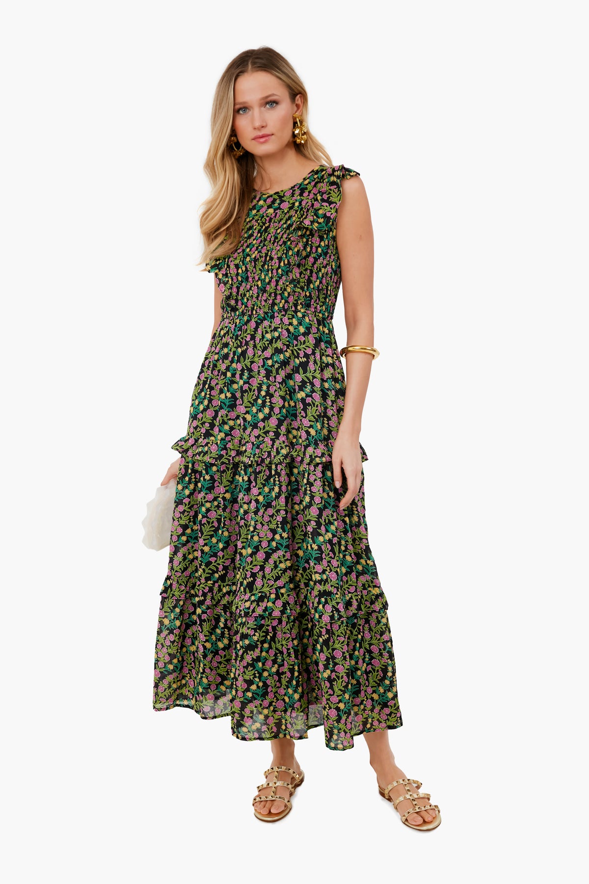 A woman in a sleeveless, ankle-length black floral Banjanan dress made of organic cotton with handmade embroidery, styled with gold jewelry, tan sandals, and a white clutch.