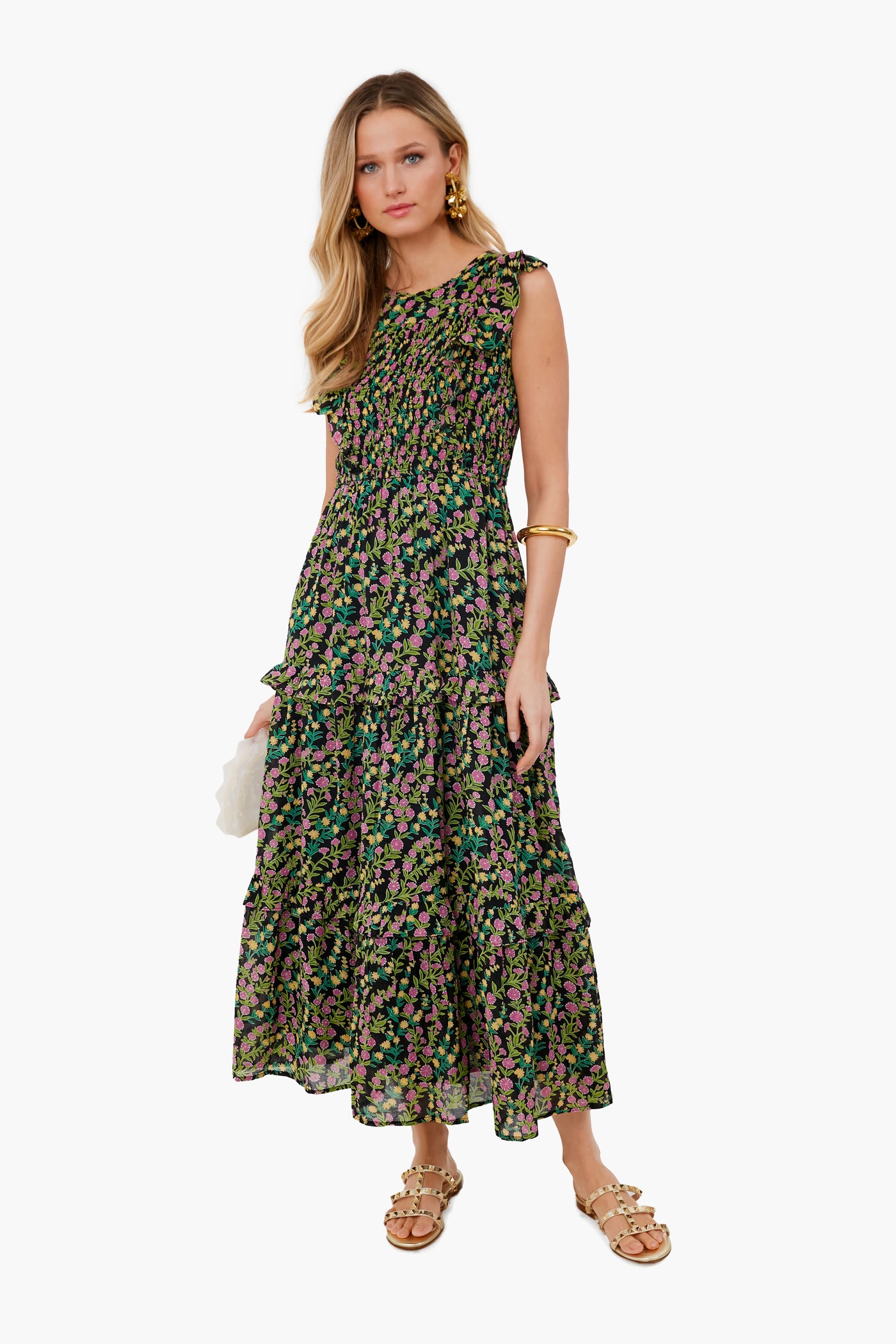 A woman in a sleeveless, ankle-length black floral Banjanan dress made of organic cotton with handmade embroidery, styled with gold jewelry, tan sandals, and a white clutch.