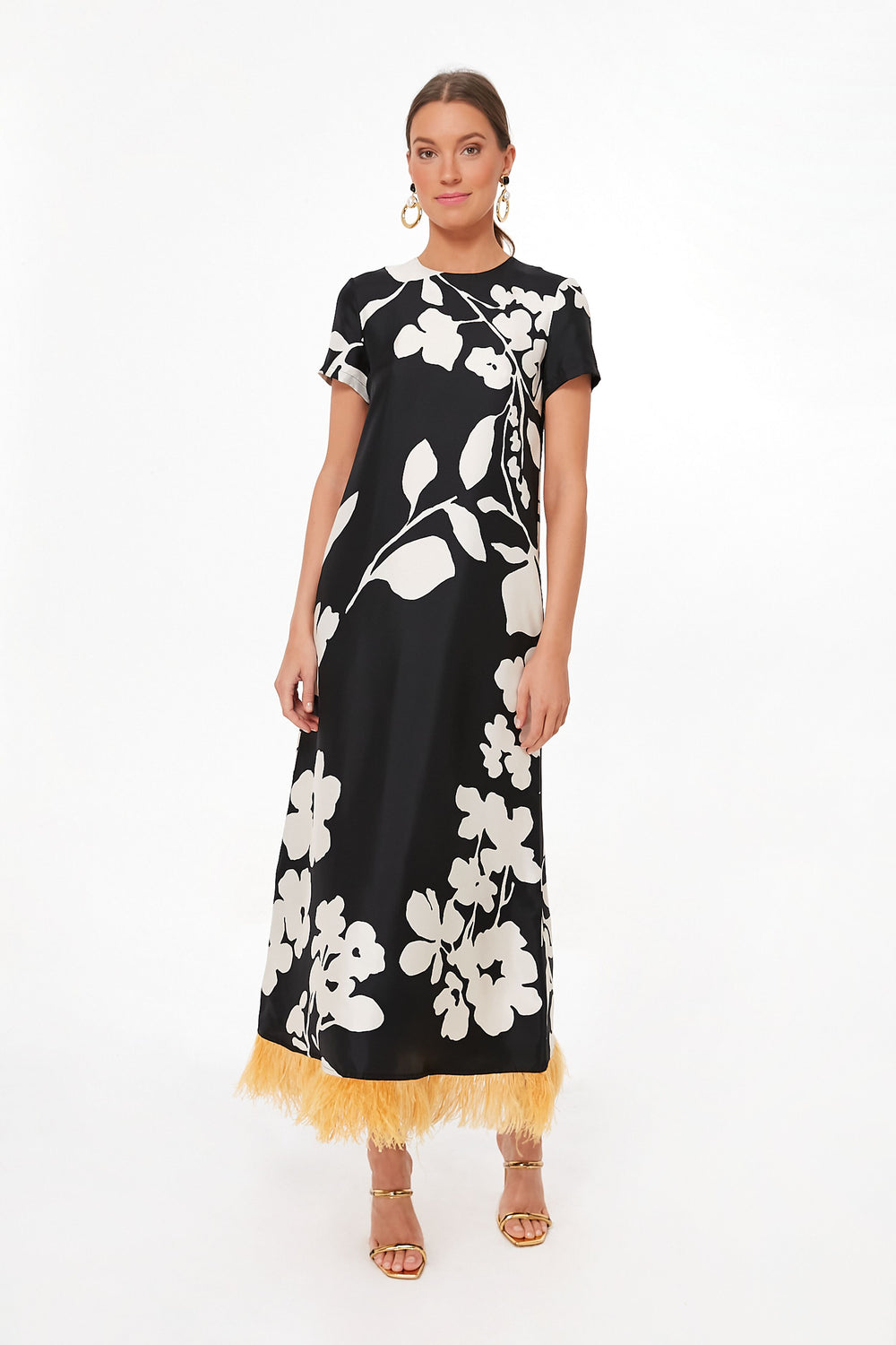 A woman wears a La DoubleJ black silk twill maxi dress with bold white floral patterns and ostrich feather trim, paired with gold hoop earrings and strappy heels.