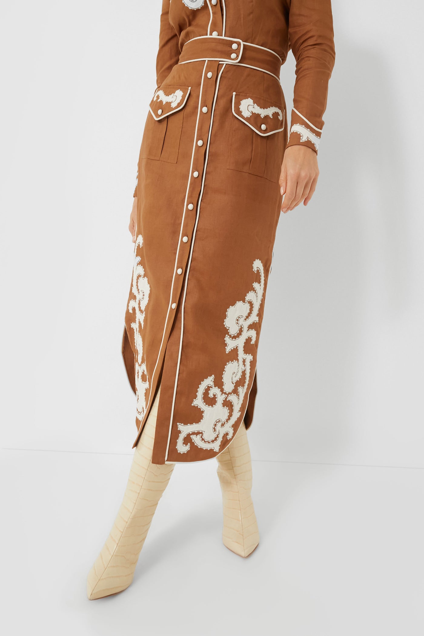 Model wears an Alemais high-waisted linen skirt with handmade applique details and a matching belt, paired with cream knee-high boots; shown from waist to feet.