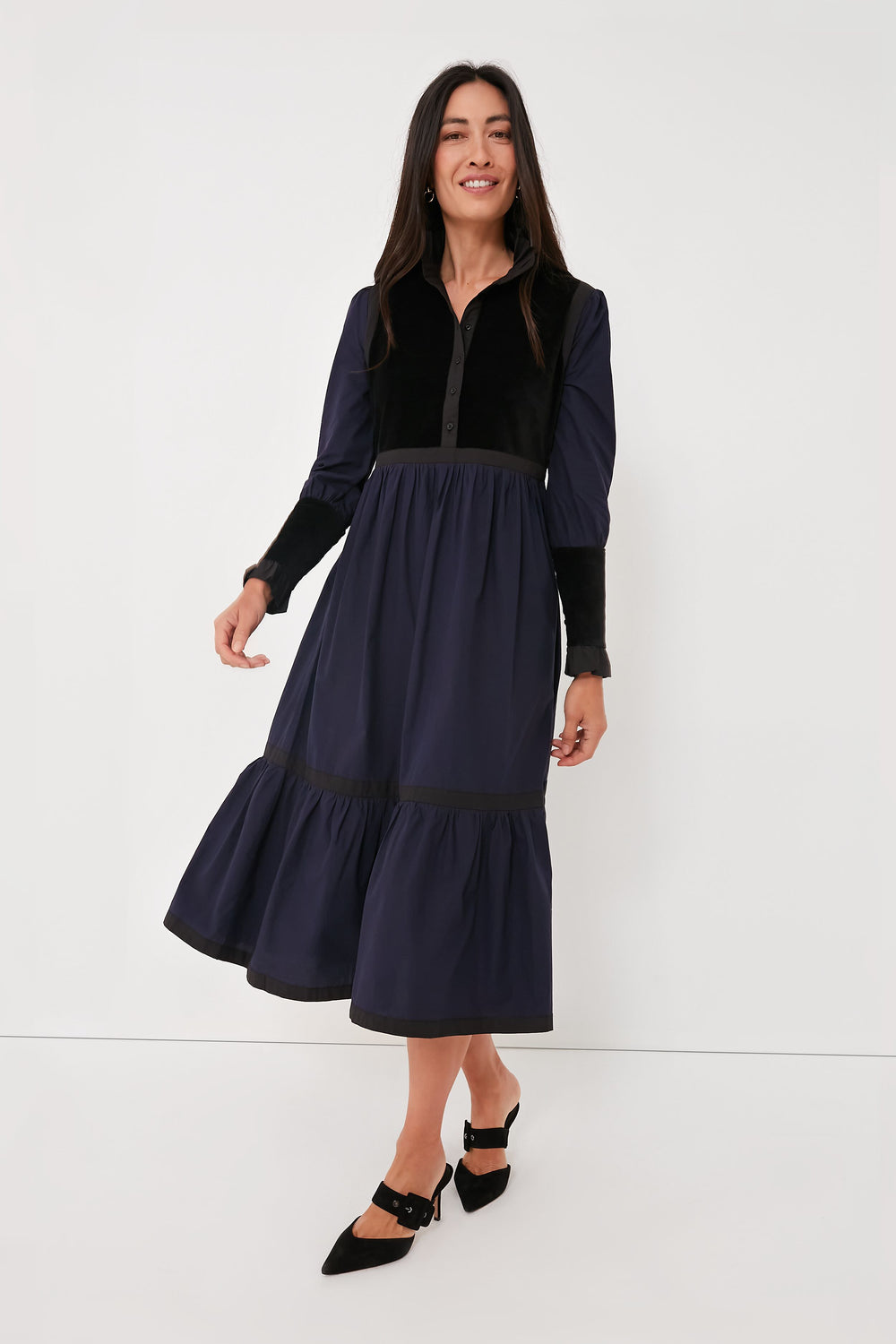 Woman in a BATSHEVA long-sleeve dress with velvet trim, showcasing a navy and black tiered midi skirt, paired with black pointed-toe shoes.