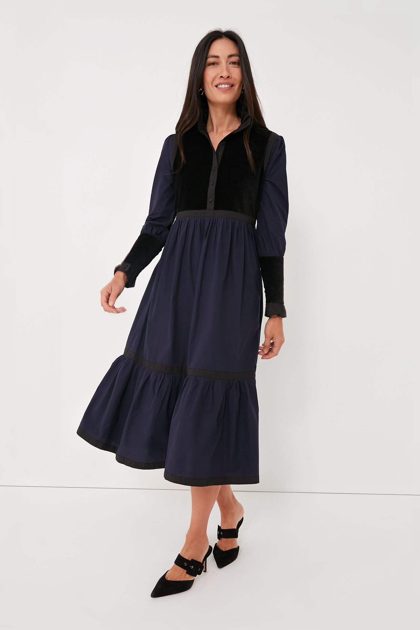 Woman in a BATSHEVA long-sleeve dress with velvet trim, showcasing a navy and black tiered midi skirt, paired with black pointed-toe shoes.