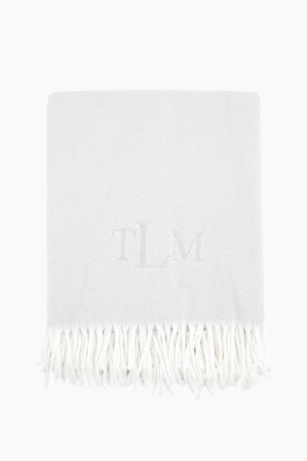 A light grey Matouk Silver Pezzo Throw features a white fringe trim and the monogram TLM embroidered at the center.