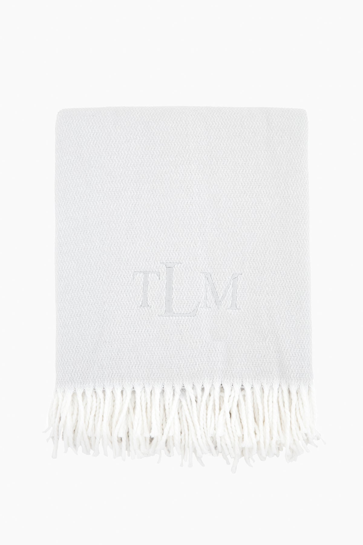 A light grey Matouk Silver Pezzo Throw features a white fringe trim and the monogram TLM embroidered at the center.